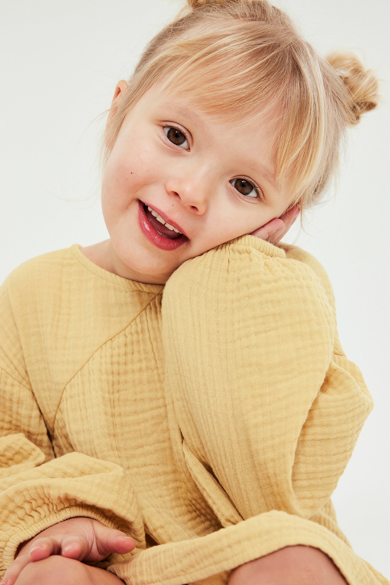 Zbliżenie uśmiechniętego dziecka w żółtej teksturowanej muślinowej sukience z bufiastymi długimi rękawami.