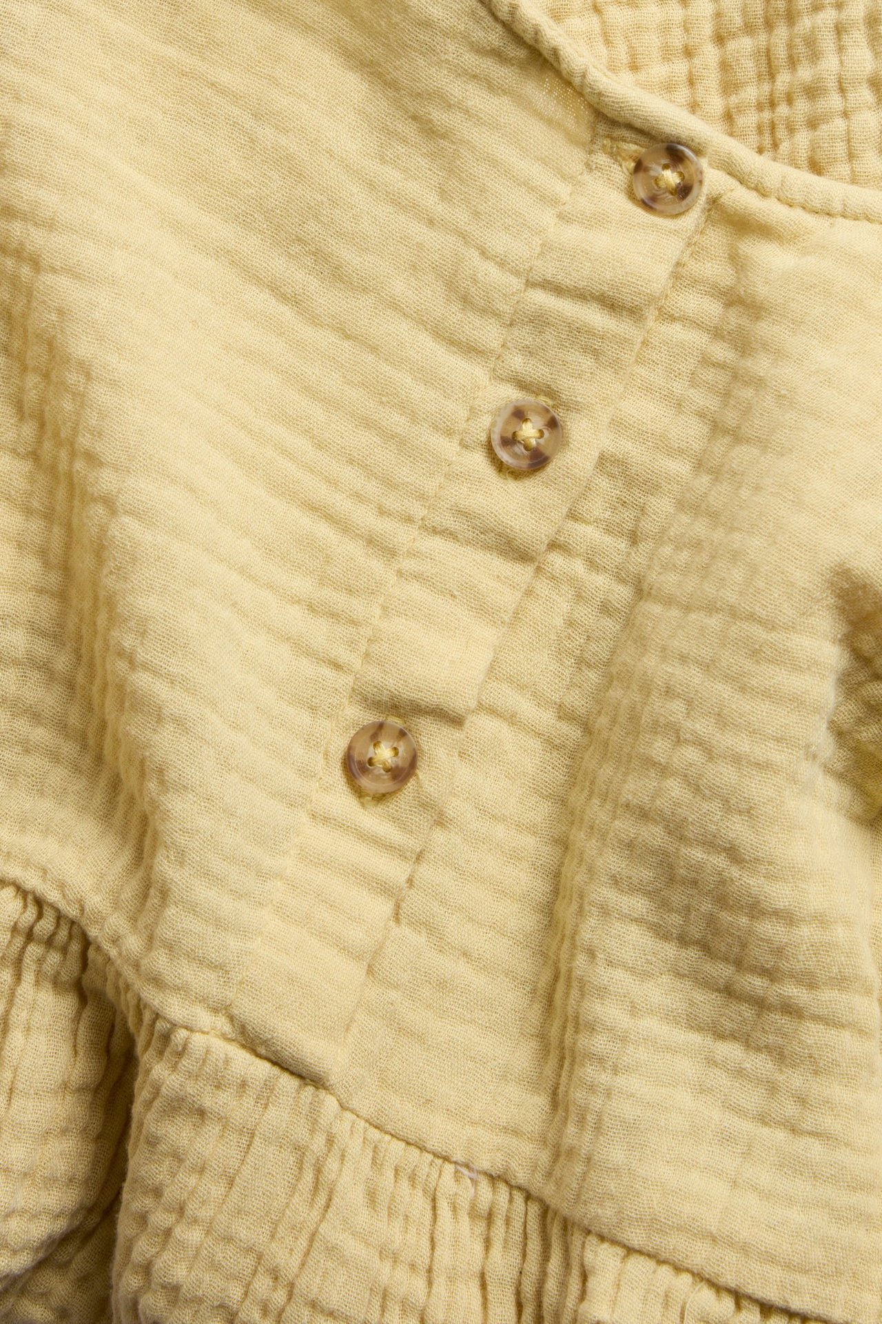 Nærbilde av en ensfarget gul muslinkjole med tre brune knapper og en rynket detalj.