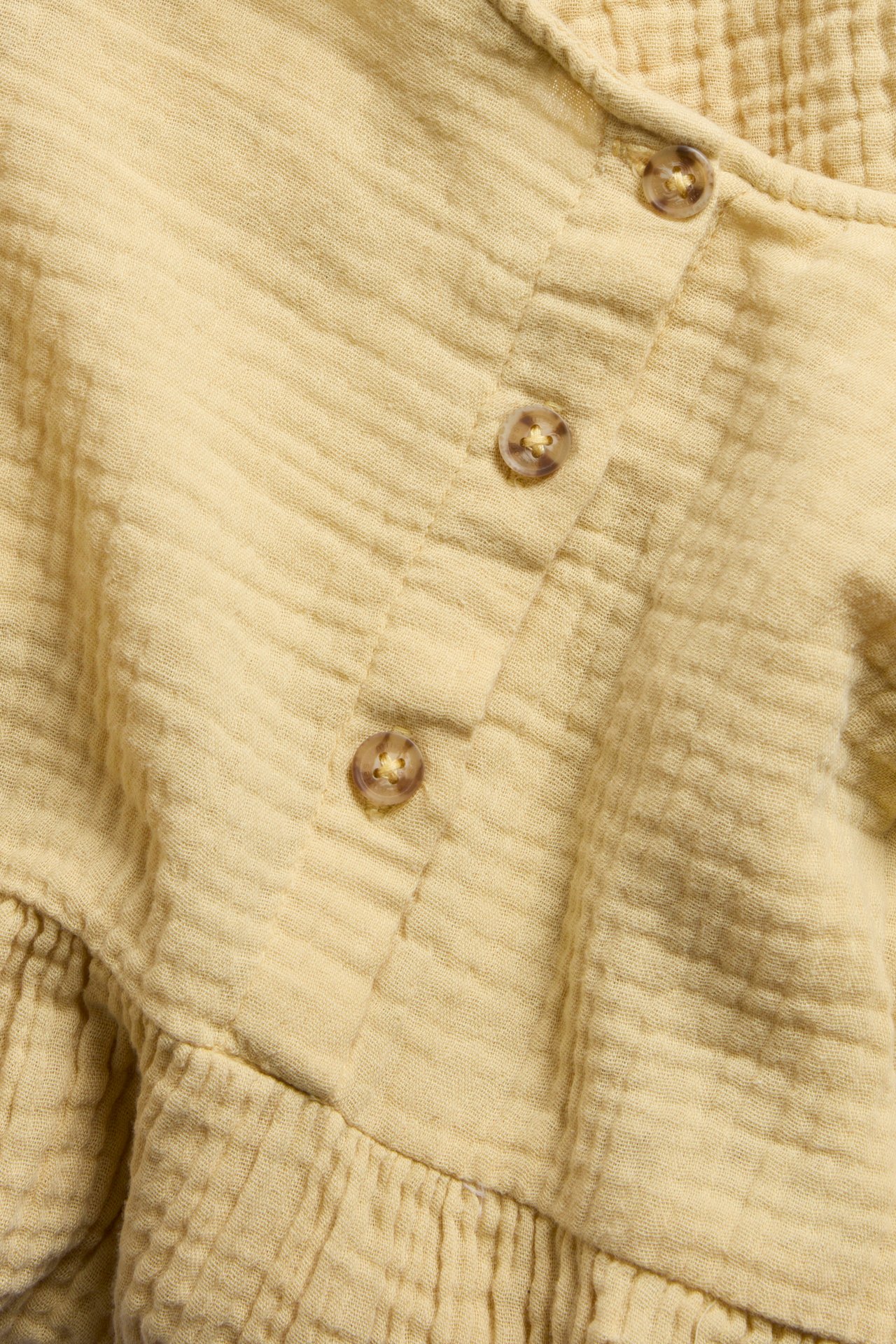 Zbliżenie żółtej muślinowej sukienki o marszczonej fakturze, z częściową listwą guzikową i marszczeniami.