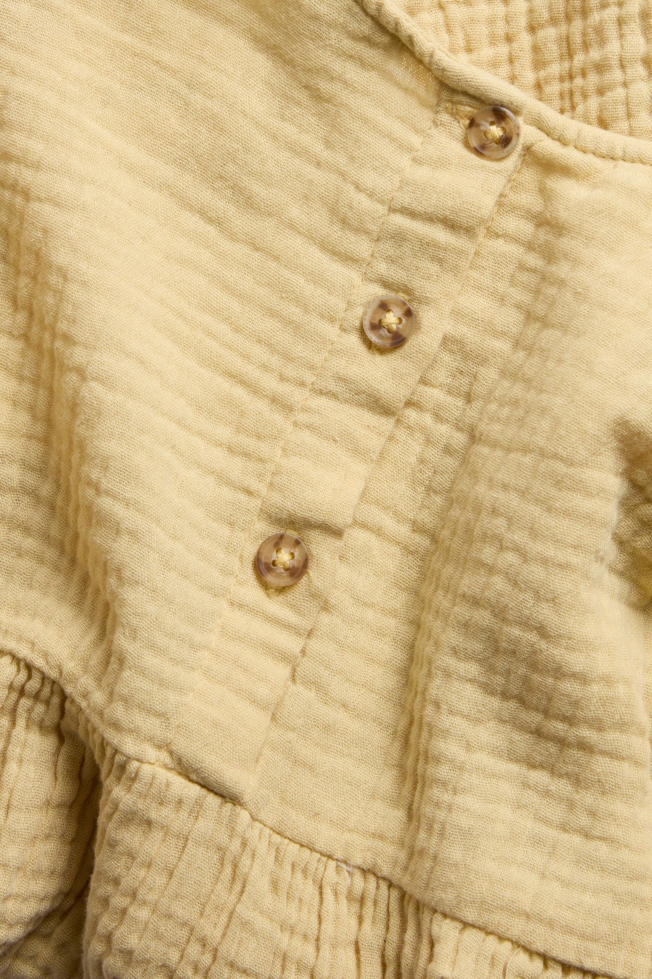 Zbliżenie żółtej muślinowej sukienki o marszczonej fakturze, z częściową listwą guzikową i marszczeniami.