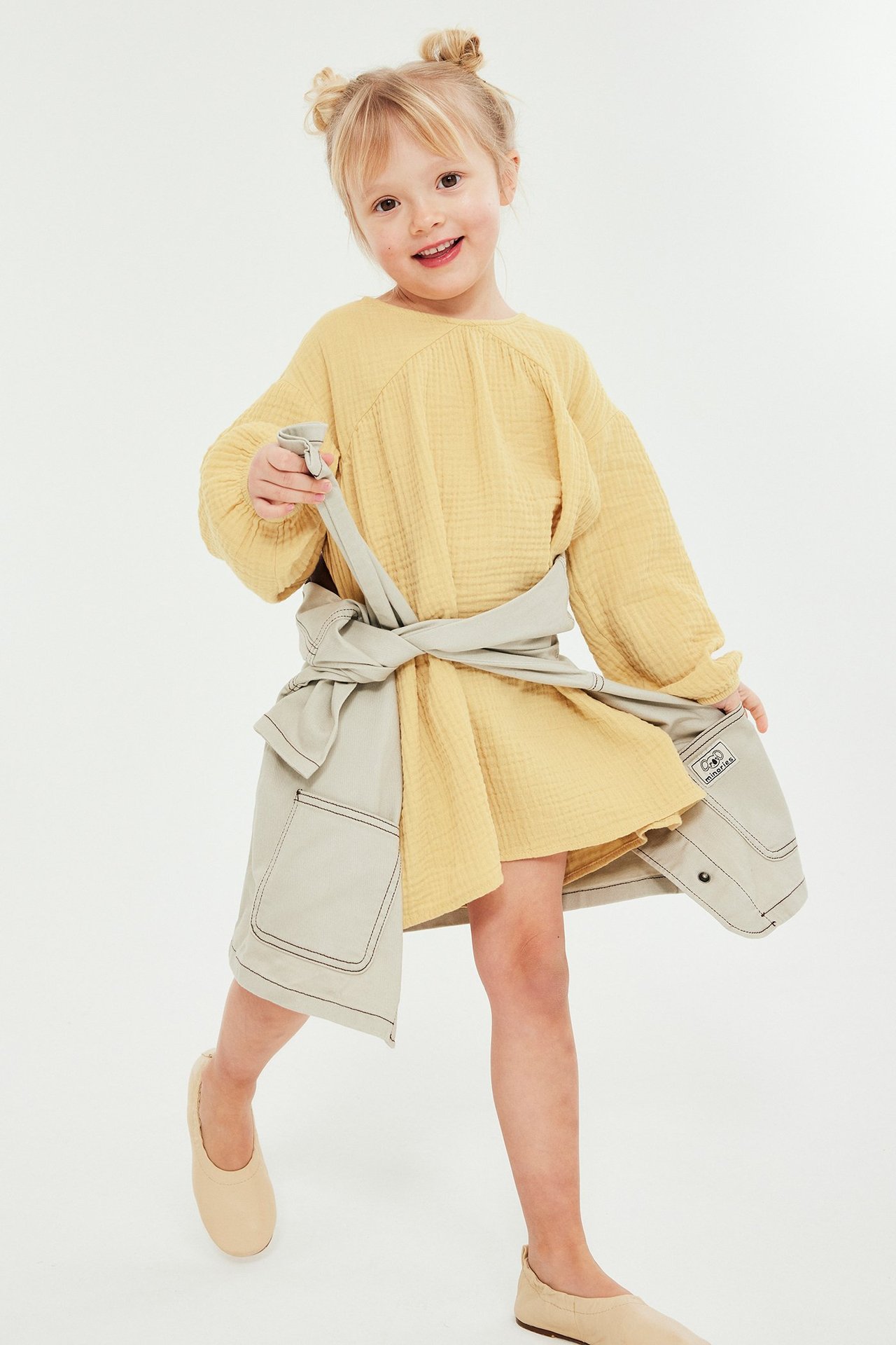 Frontbilde av et barn i gul muslin-kjole, med lyst beige omslagsskjørt og beige ballerinasko.