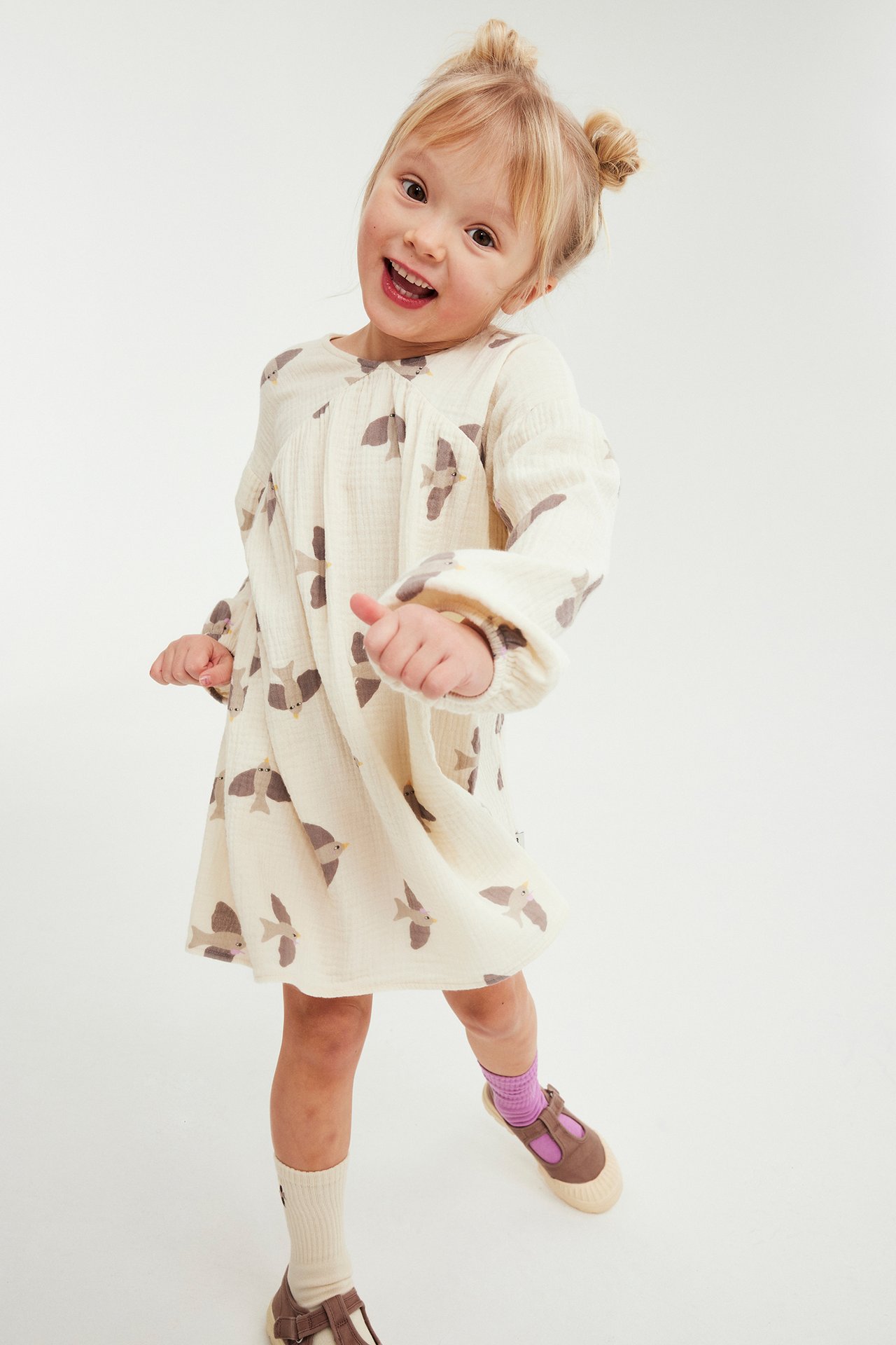 Uśmiechnięte dziecko w luźnej beżowej sukience z muślinu w ptaki, różnych skarpetkach i brązowych Mary Jane.