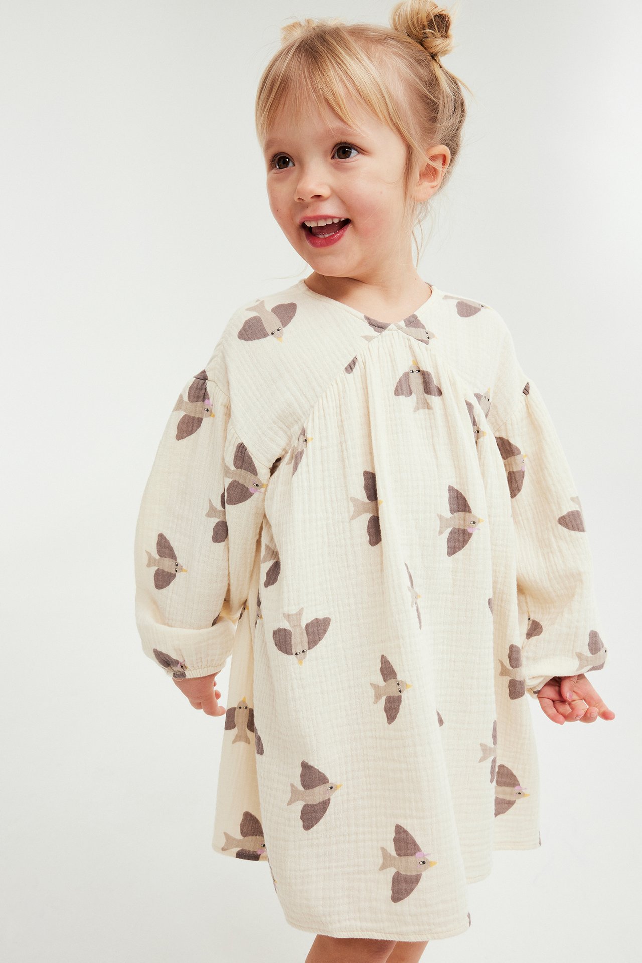 Uśmiechnięte dziecko w luźnej beżowej sukience muślinowej z długimi rękawami i brązowymi ptakami.