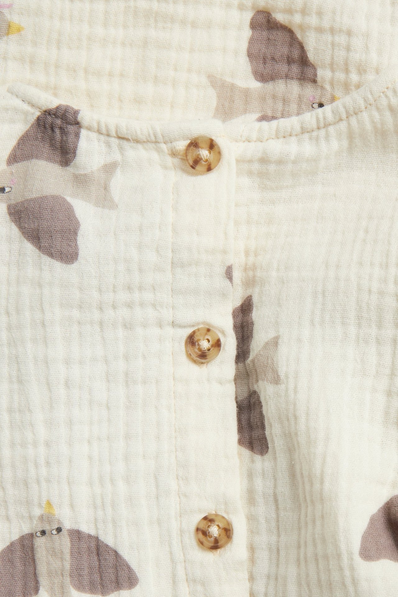 Zbliżenie na beżową muślinową sukienkę dziecięcą z brązowym wzorem ptaków i guzikami.