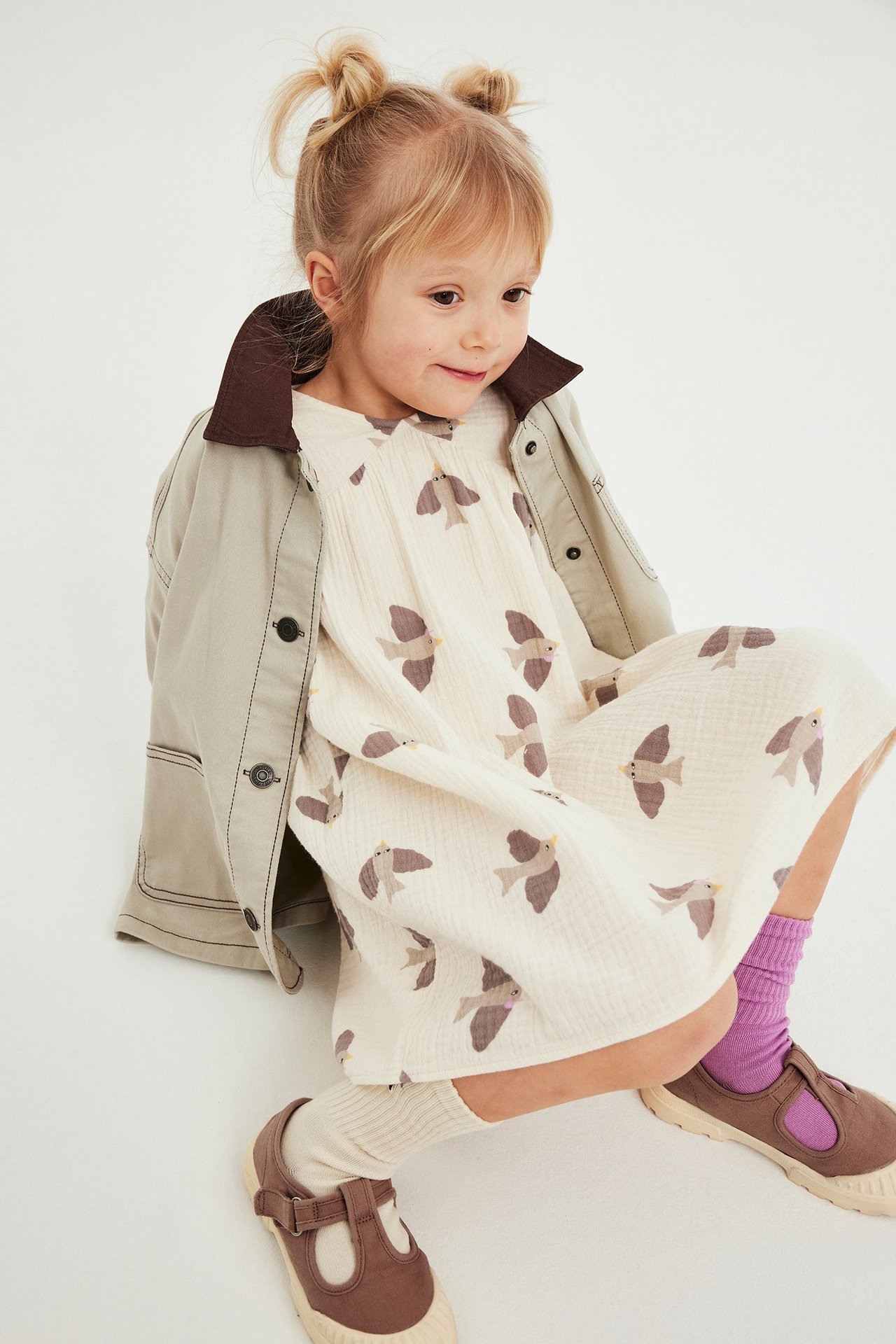 Forfra: Barn i beige fuglemønstret musselin kjole, med lys beige jakke og tofargede sokker.