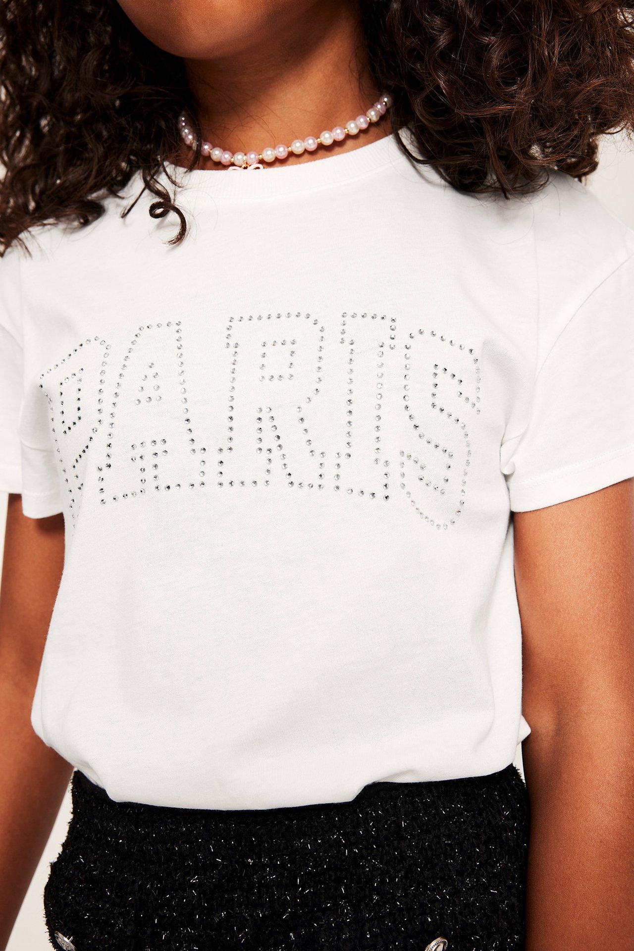 Nærbilde forfra, barn i offwhite bomulls-T-skjorte med "PARIS"-trykk i rhinestones, rosa perlekjede.