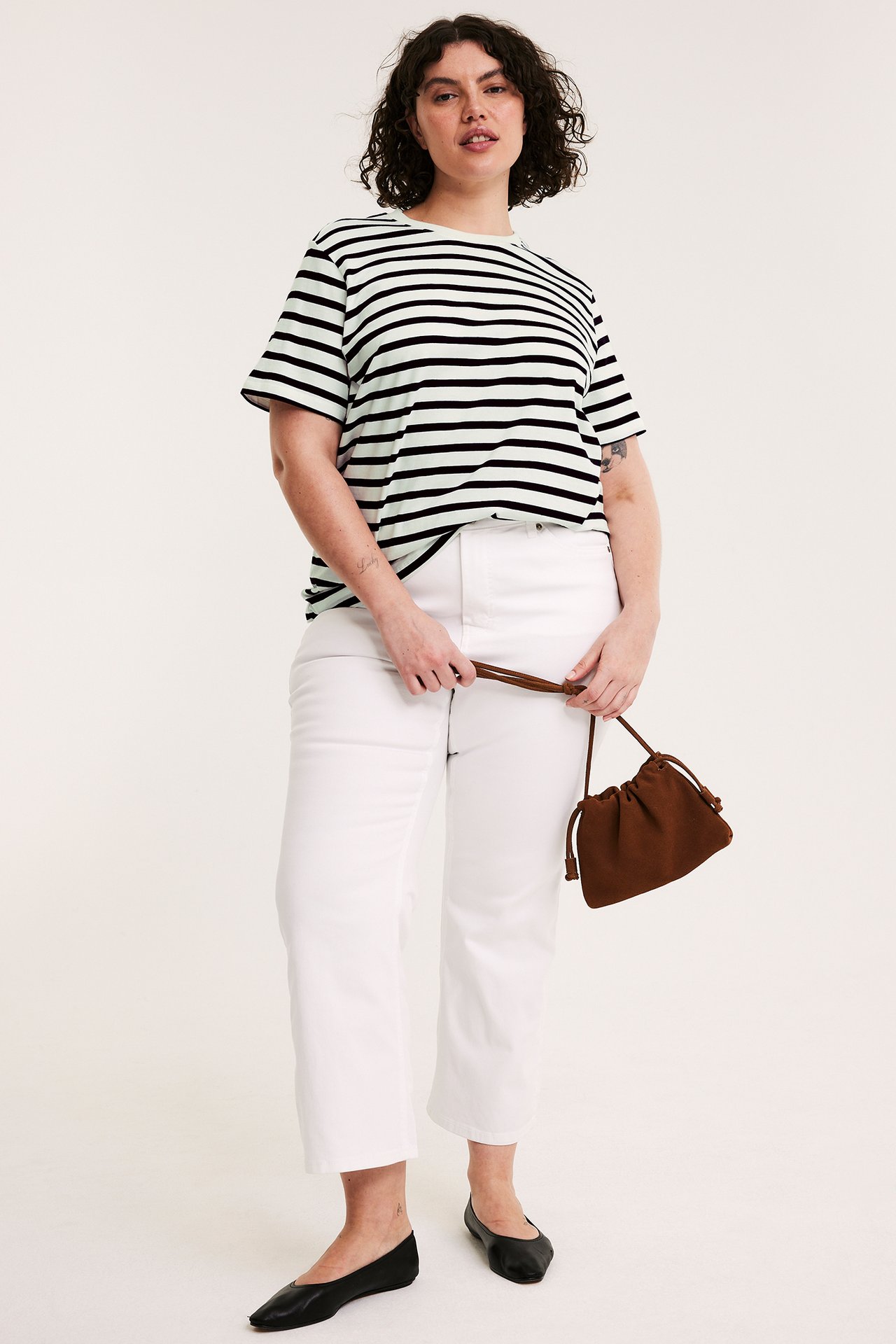 Framifrån: Kvinna i ljusgrön/svartrandig bomulls-t-shirt, vita jeans, svarta ballerinaskor, håller brun väska.