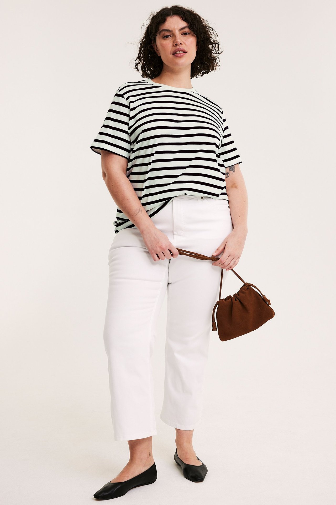 Edestä: Nainen yllään vaaleanvihreä/mustaraitainen puuvilla-t-paita, valkoiset farkut, mustat ballerinat, pitäen ruskeaa laukkua.