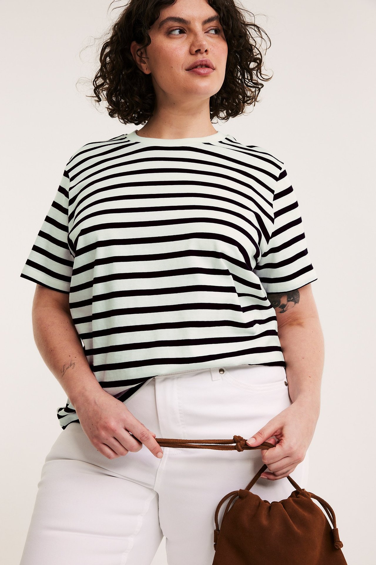 Edestä: Nainen yllään musta- ja vaaleanvihreäraitainen puuvillapaita, valkoiset housut, pitäen ruskeaa laukkua.