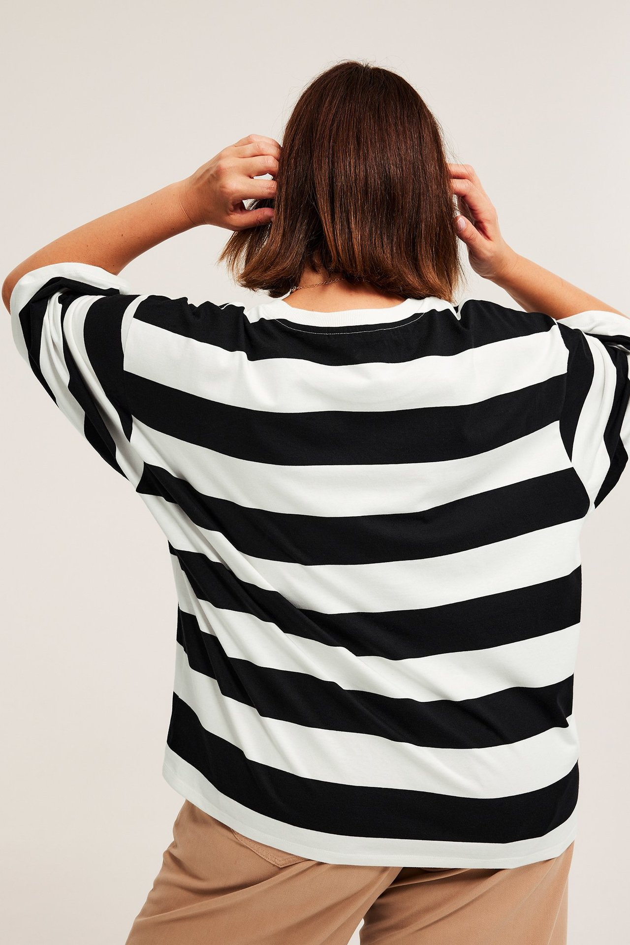 Widok z tyłu kobiety w czarno-białej, paskowanej, oversize bawełnianej koszulce i beżowych spodniach.