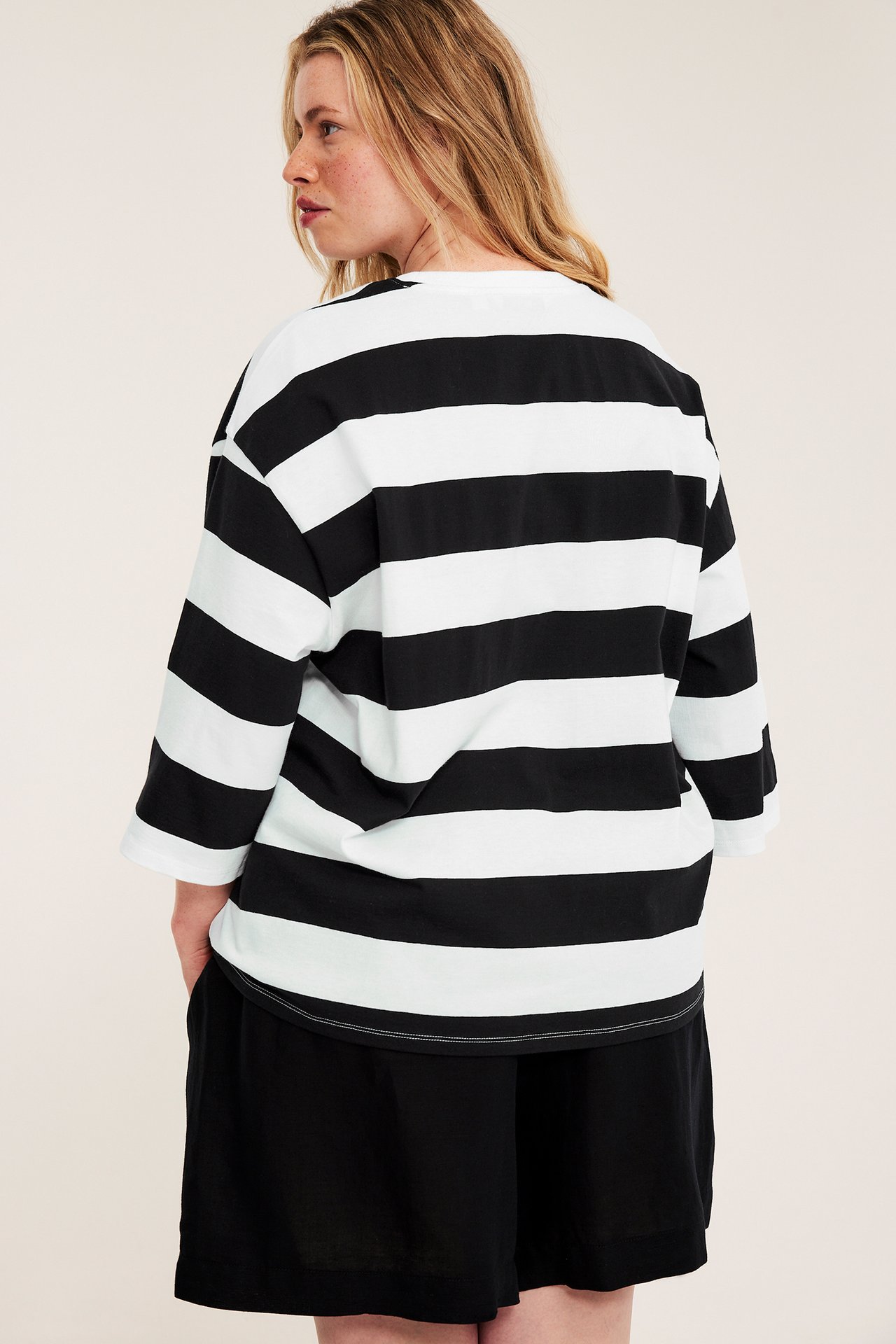 Bakfra-sidevisning av kvinne i oversized svart- og hvitstripet t-skjorte med formede ermer og svarte shorts.