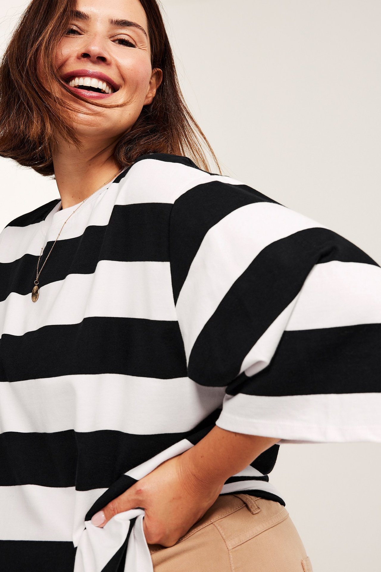 Uśmiechnięta kobieta w oversize'owym czarno-białym T-shircie w paski, beżowych spodniach i złotym naszyjniku.