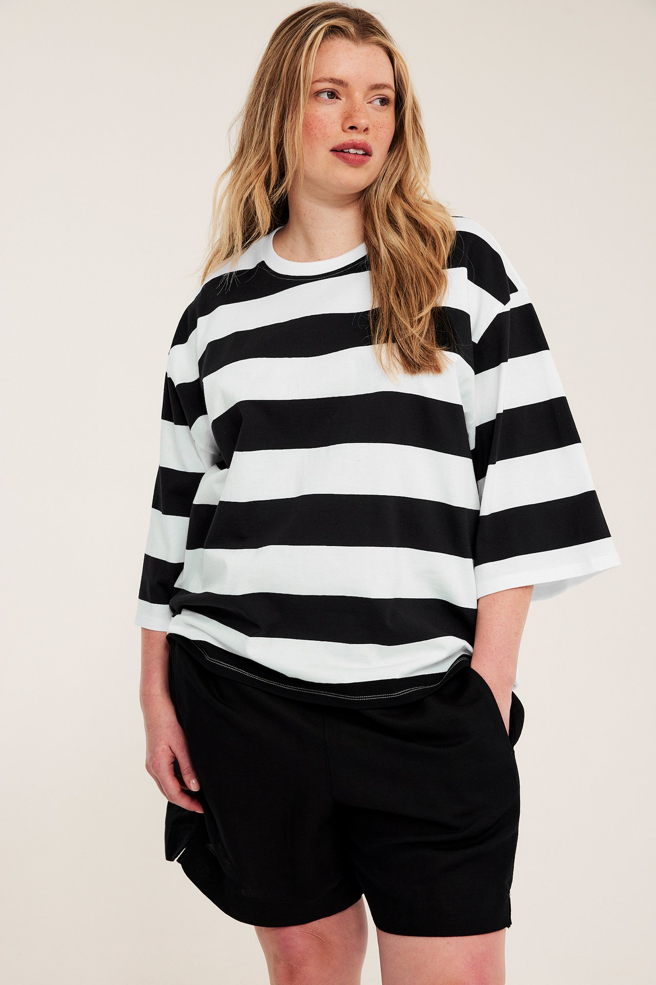 Frontvisning, kvinne i offwhite og svartstripet oversize bomull T-skjorte med svarte shorts.