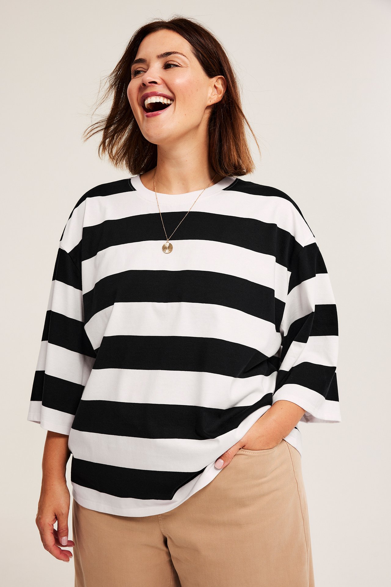Z przodu, uśmiechnięta kobieta w luźnej koszulce w czarno-białe paski, beżowych spodniach, złotym naszyjniku.