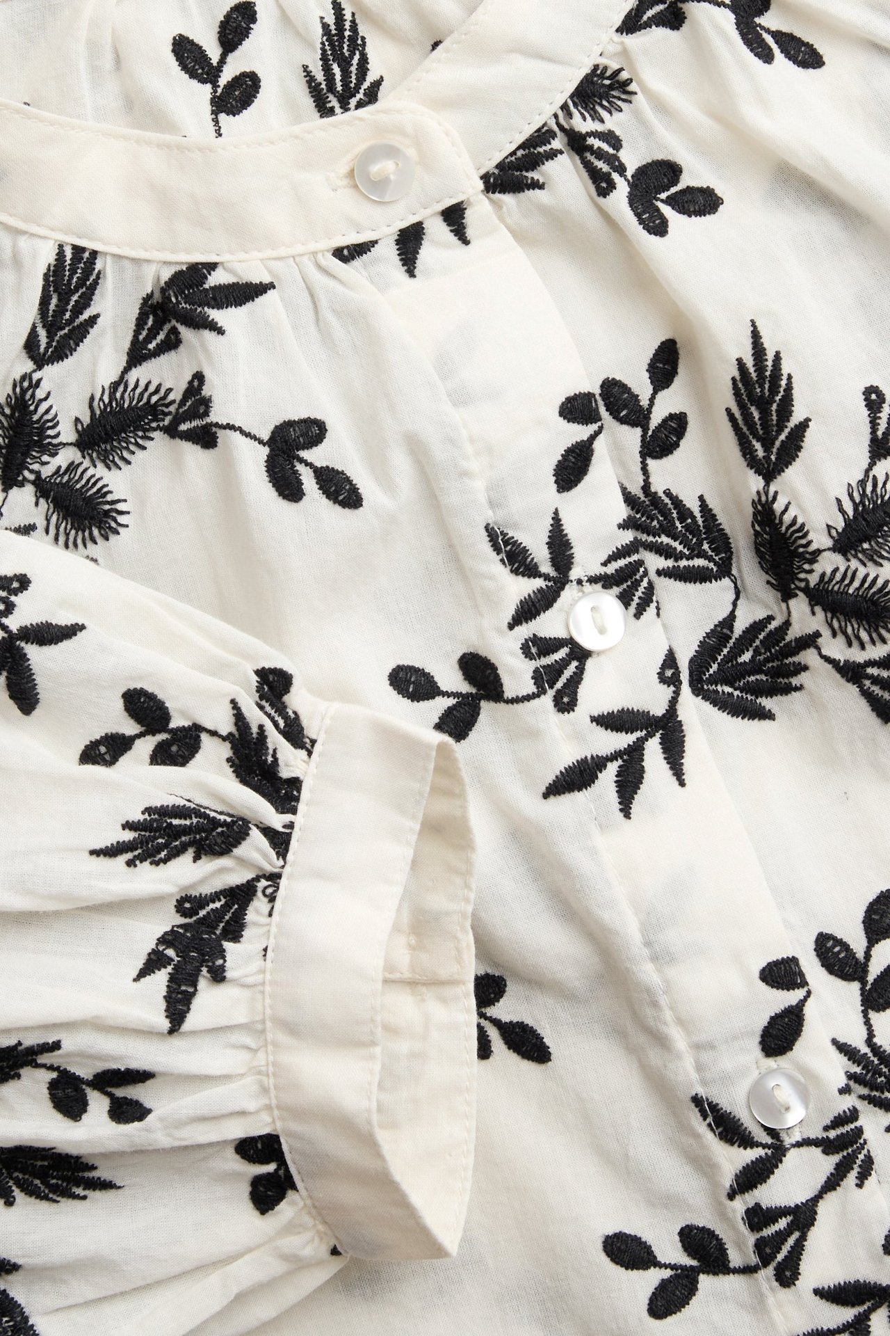 Zbliżenie na ecru bawełnianą bluzkę z czarnym haftem kwiatowym, guzikami i marszczeniami.