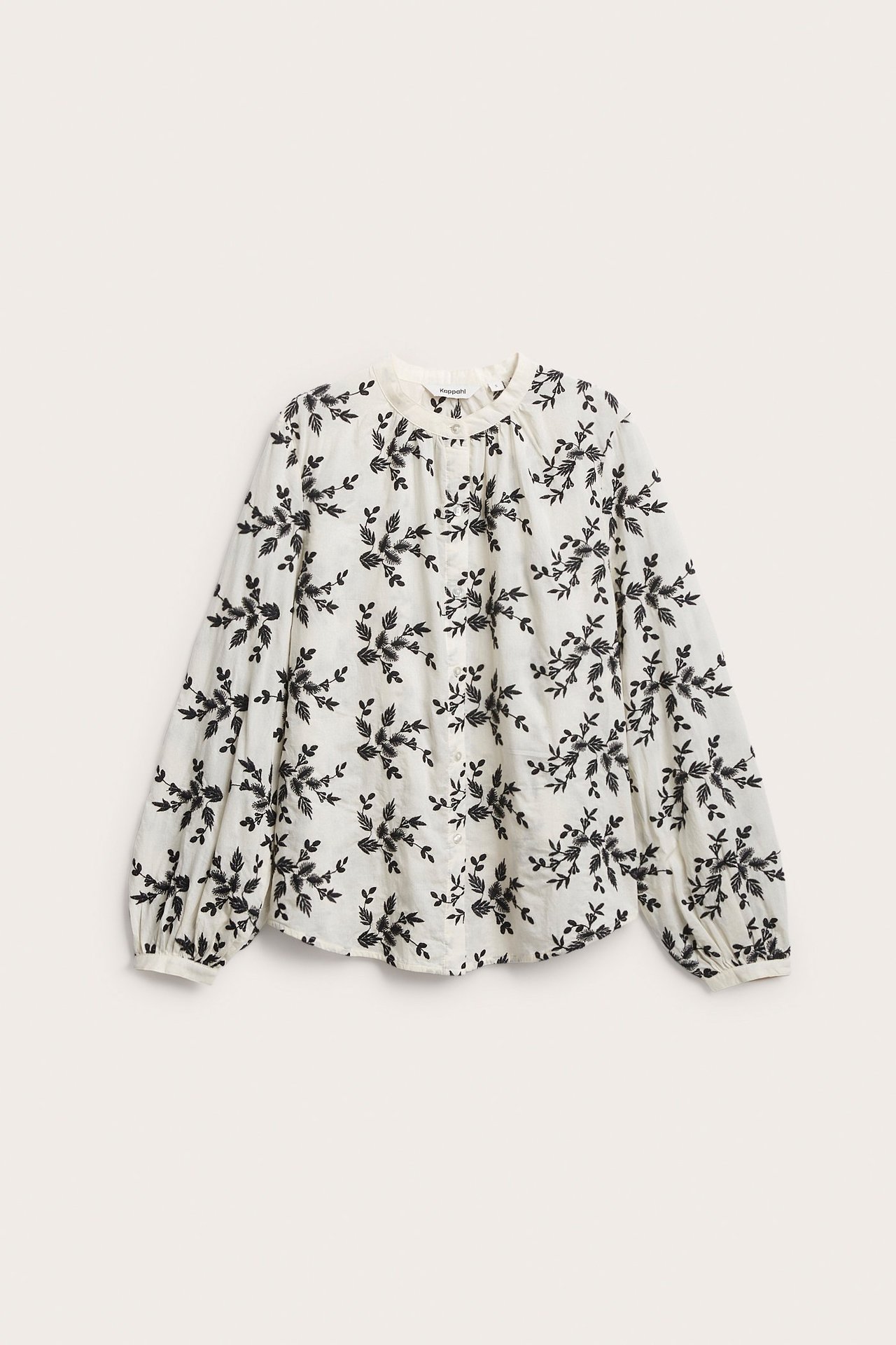 Frontvisning av en offwhite bomullsbluse med svart brodert blomstermønster og knapper foran.