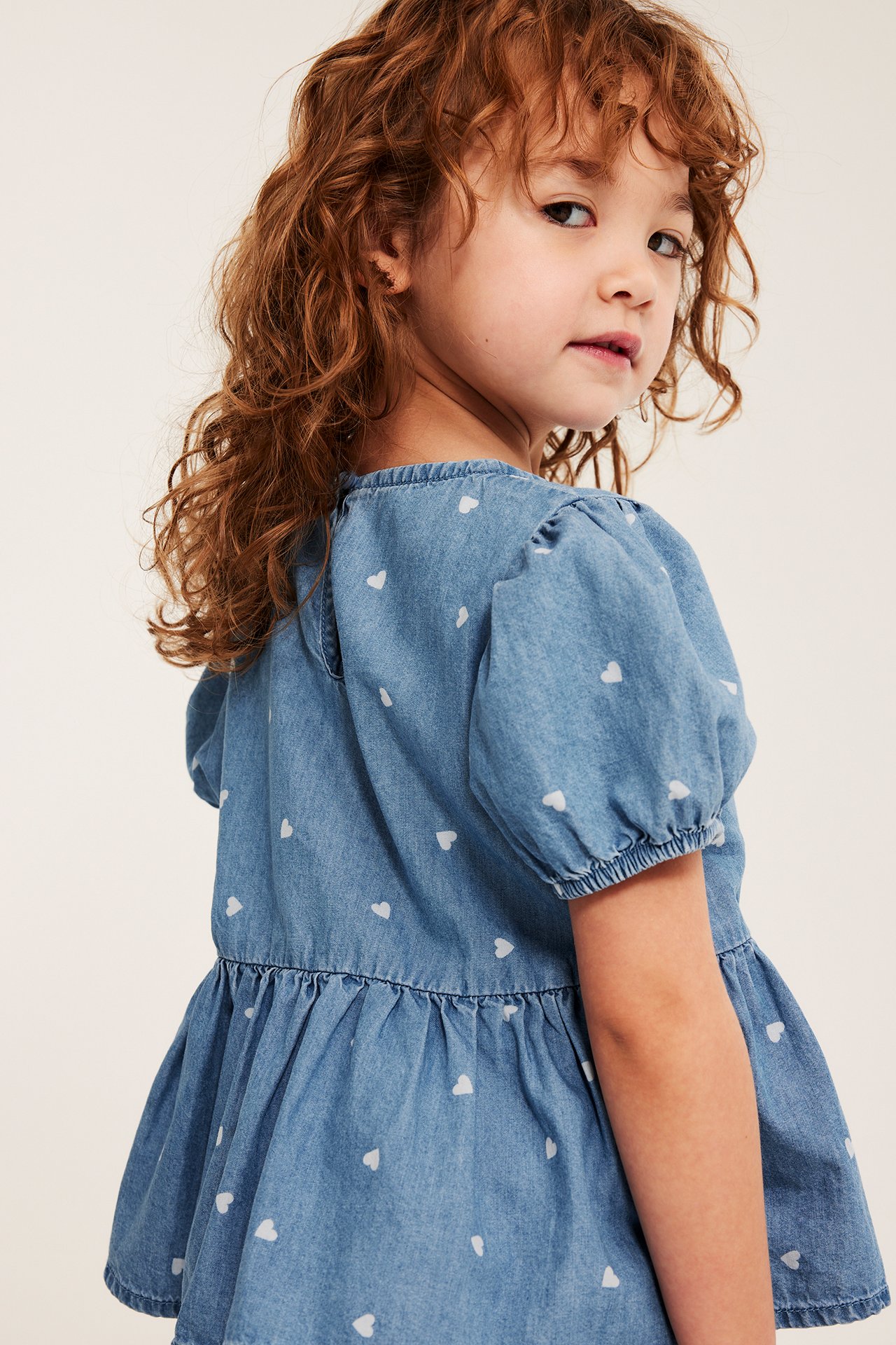 Baksidebilde av et barn i en lys denimbluse med hvitt hjerteprint, puffermer og rynket midje.