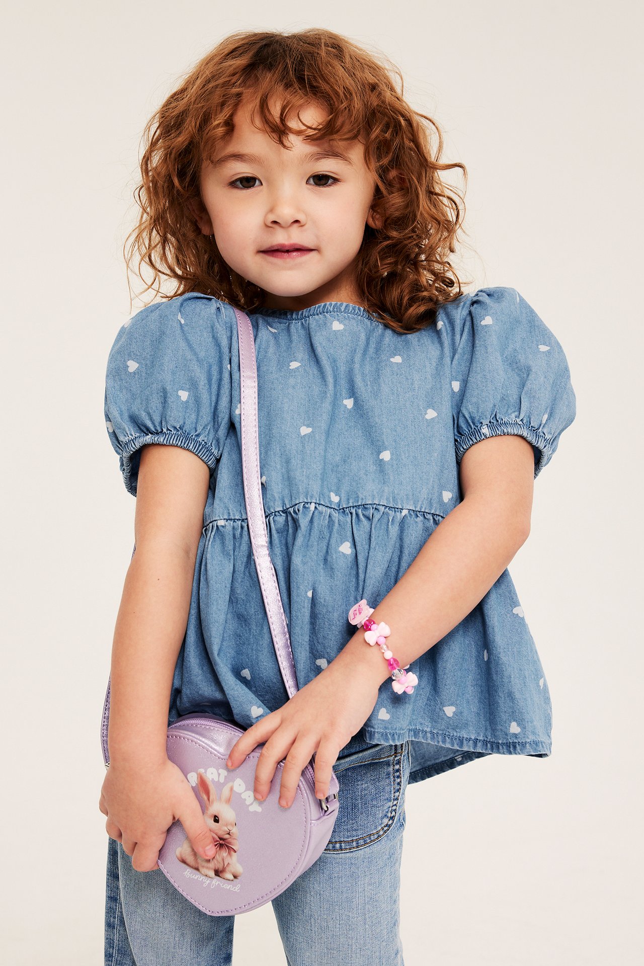 Front: Barn i lys dongeribluse med hjertemønster, lyseblå jeans, holder lilla hjerteveske og rosa armbånd.