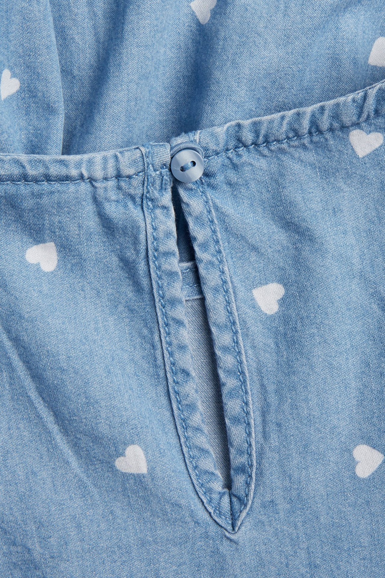 Zbliżenie na dziecięcą jasnoniebieską bluzkę dżinsową z białymi sercami i zapięciem na guzik z tyłu.