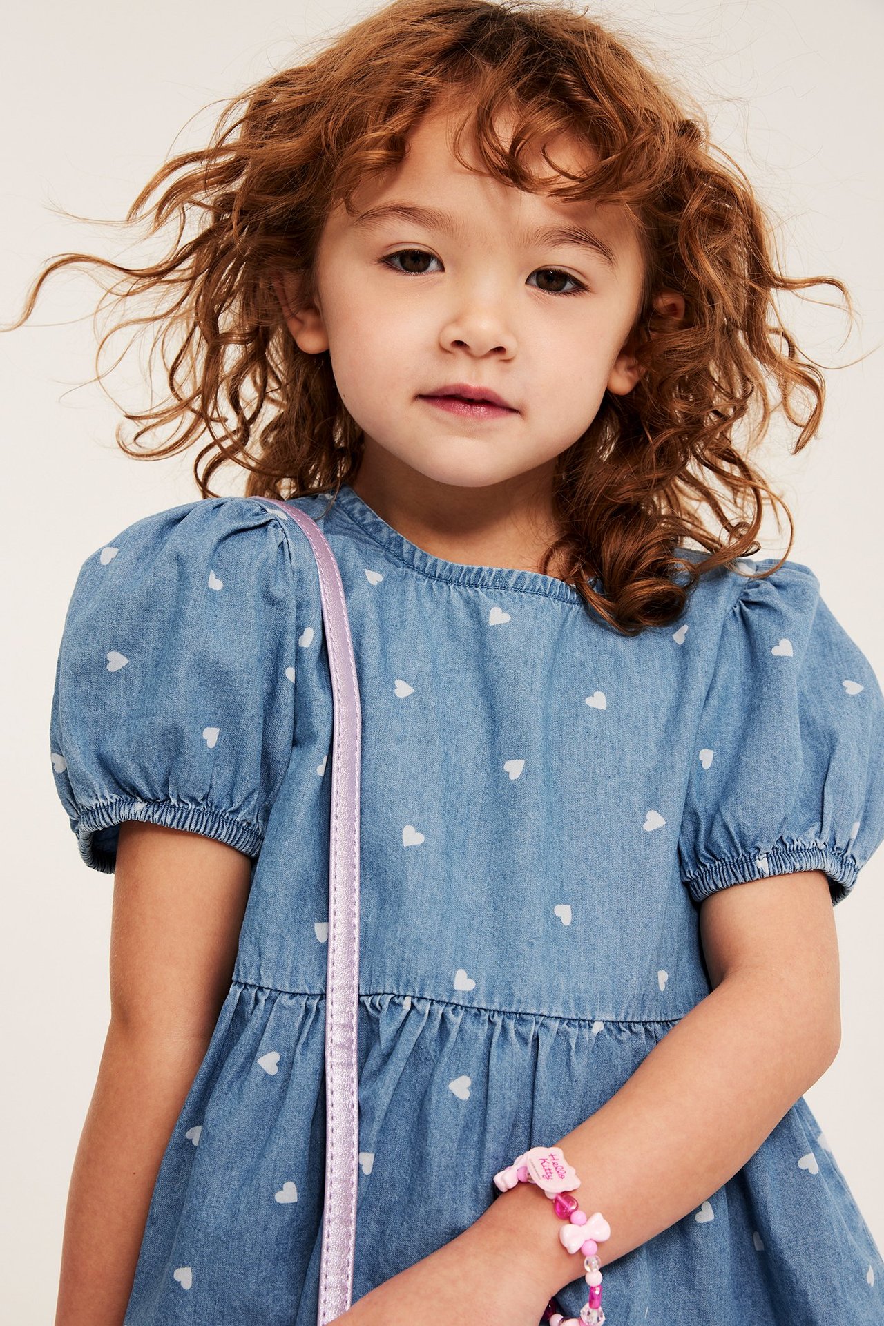 Framifrån: ett barn i ljusblå hjärtmönstrad denimblus med puffärm och ett rosa pärlarmband.