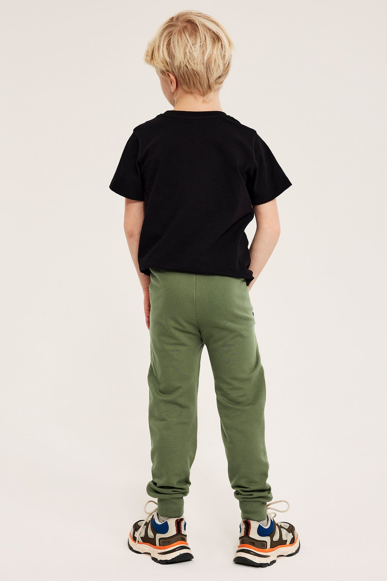 Barn, bakfra: mørkegrønn joggebukse med forsterkede knær, ribbekanter og svart t-skjorte.