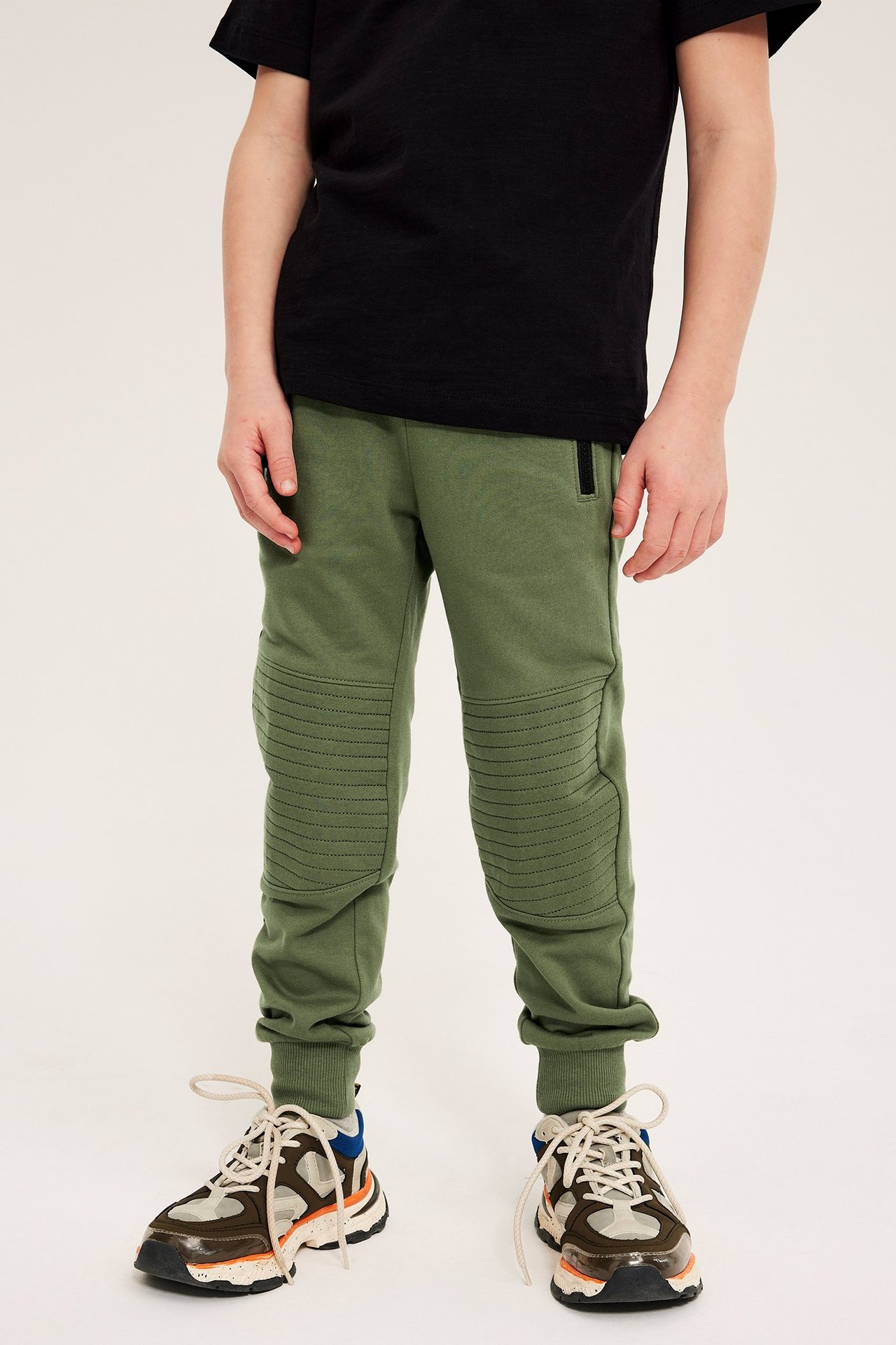 Barn, front: mørkegrønne bomulls-joggebukser med forsterkede knær, svart t-skjorte og flerfargede sko.