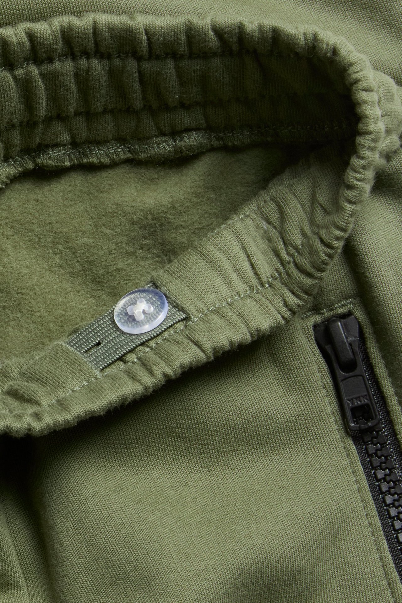 Nærbilde av mørkegrønn bomulls-joggebukse med ribbet linning, justerbar knapp og svart glidelåslomme.