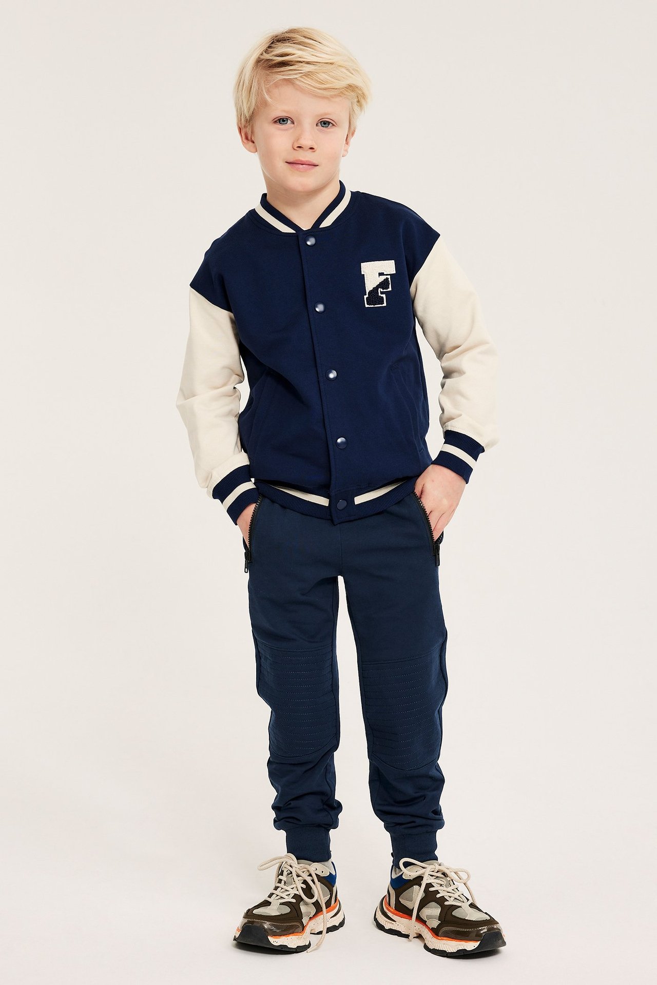 Frontvisning av et barn i mørkeblå joggebukse med forsterkede knær, en marineblå collegejakke og joggesko.