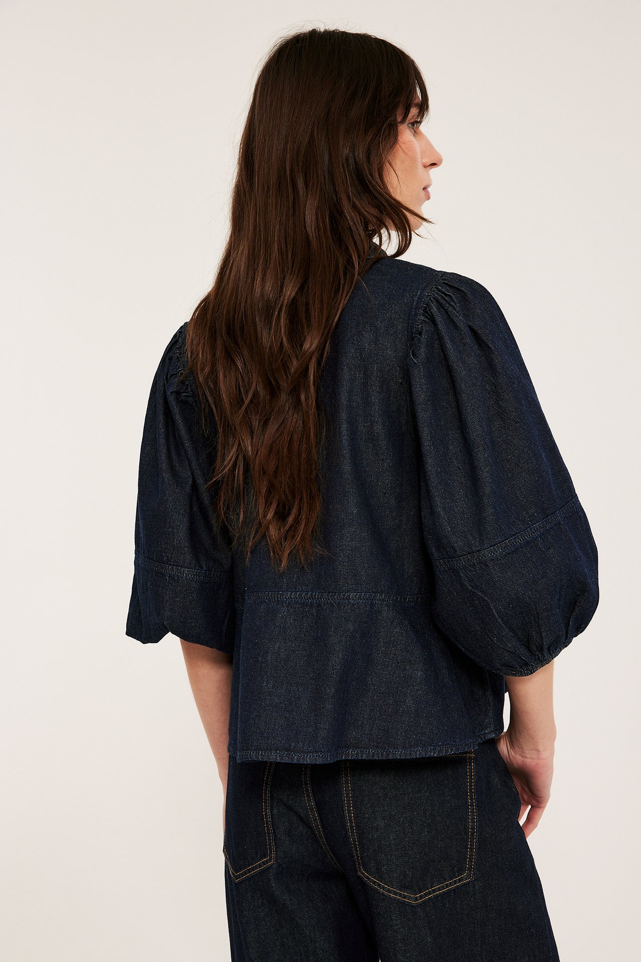 Widok z tyłu: kobieta w ciemnej dżinsowej bluzce z baskinką, bufiastymi rękawami 3/4, i dżinsach.