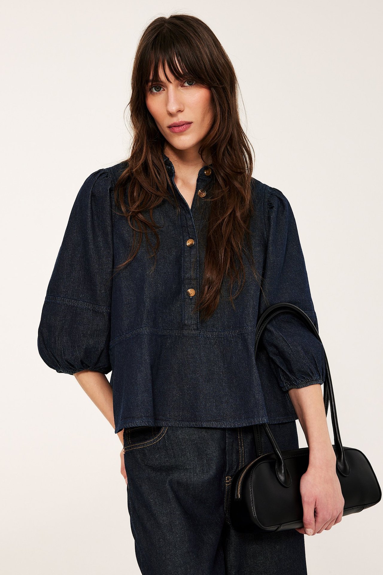 Widok z przodu: Kobieta w ciemnej jeansowej bluzce peplum z bufiastymi rękawami 3/4, jeansach i czarną torebką.