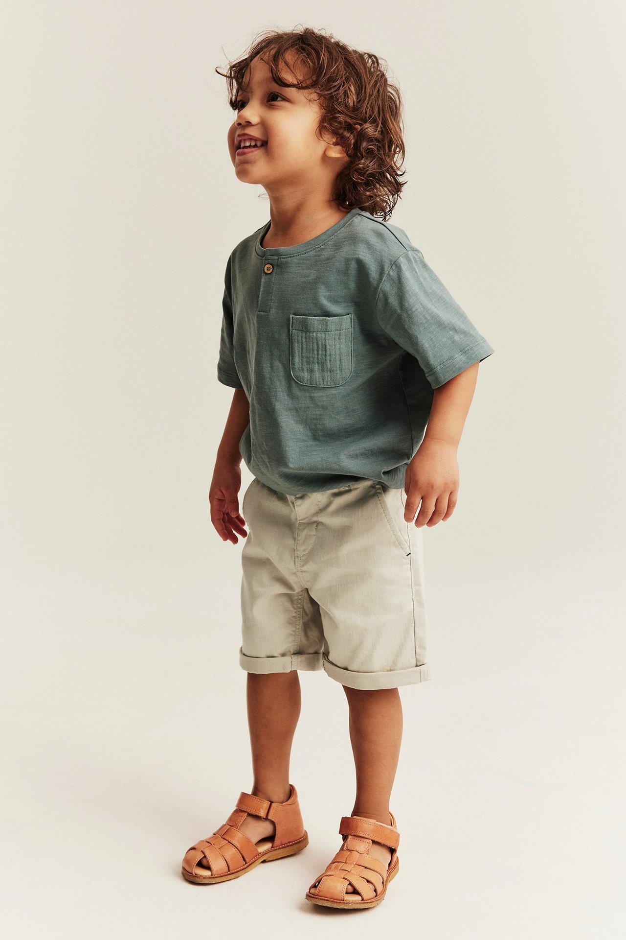 Sidevisning av et barn i beige vevde shorts med opprullede ben, grønn t-skjorte og brune sandaler.