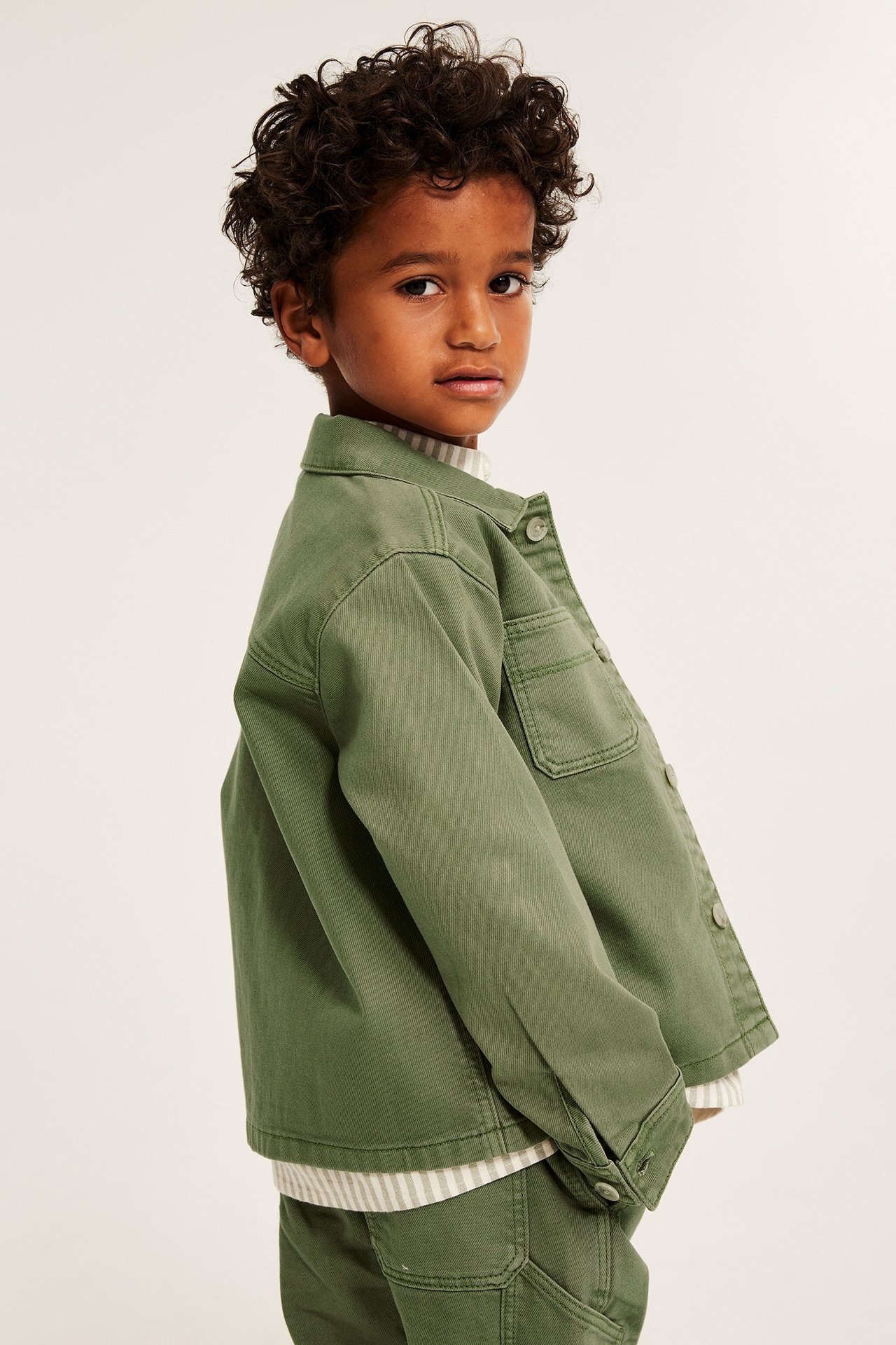 Sidovy av ett barn i mörkgrön twill-overshirt med knappar, buren över en randig topp.