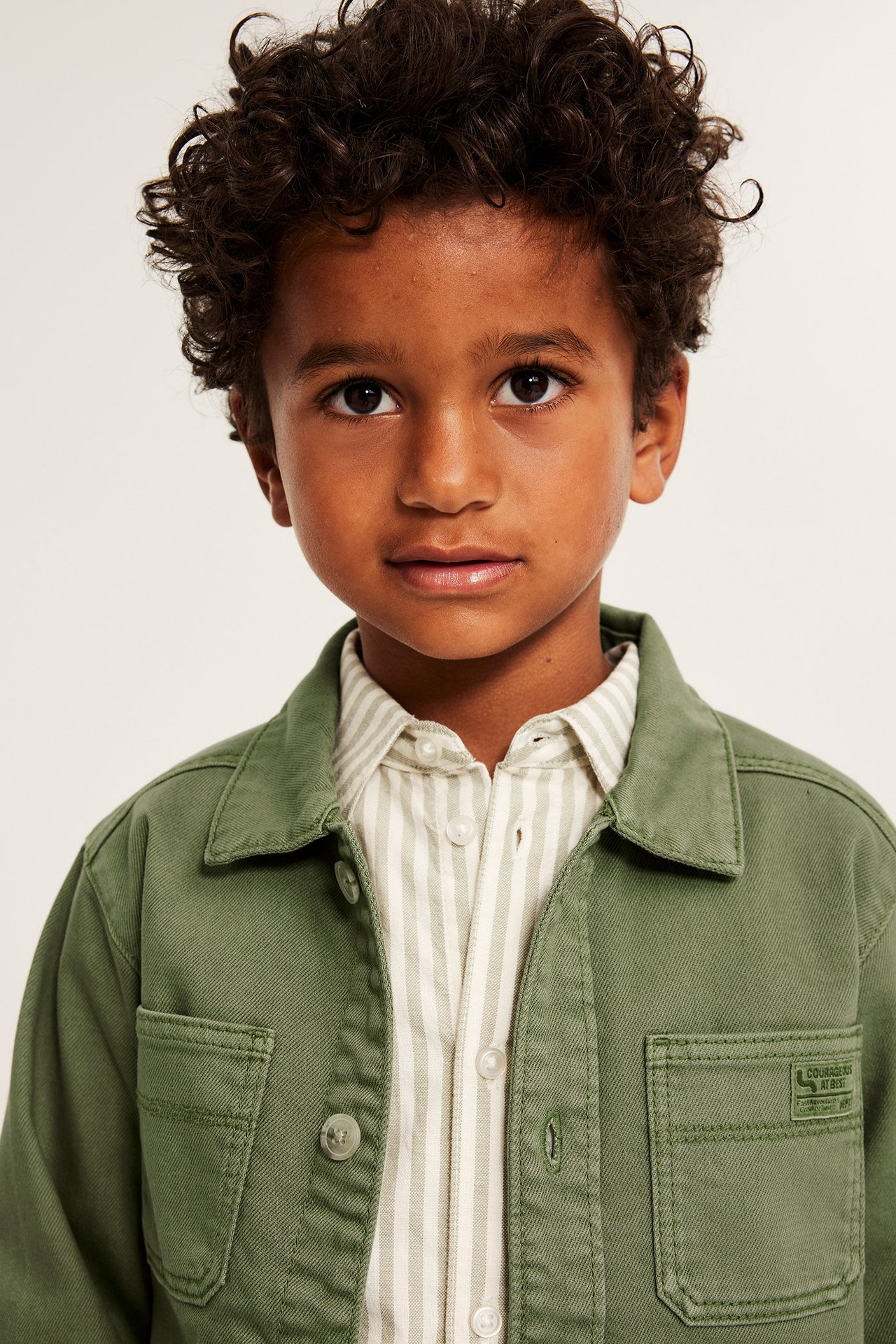 Framifrån, ett barn i mörkgrön bomullstwill-overshirt med två fickor över en ljusrandig skjorta.