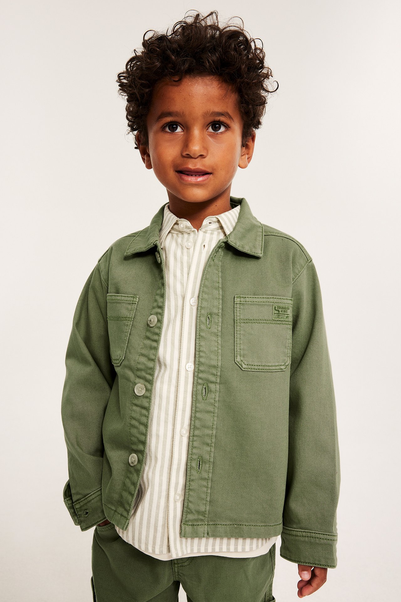 Widok z przodu: dziecko w ciemnozielonej overshirt z kieszeniami, na paskowanej koszuli i zielonych spodniach.
