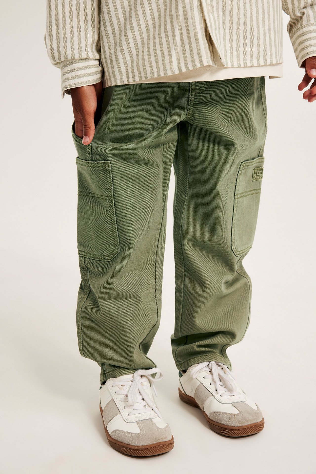 Edestä näkymä lapsesta tummanvihreissä puuvillakargohousuissa, raidallisessa paidassa ja valkoisissa-harmaissa tennareissa.