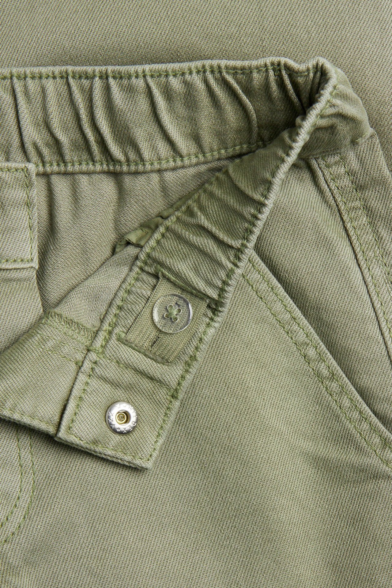 Zbliżenie ciemnozielonych bawełnianych spodni cargo, pokazujące elastyczny pas z zatrzaskiem i regulowanym guzikiem.