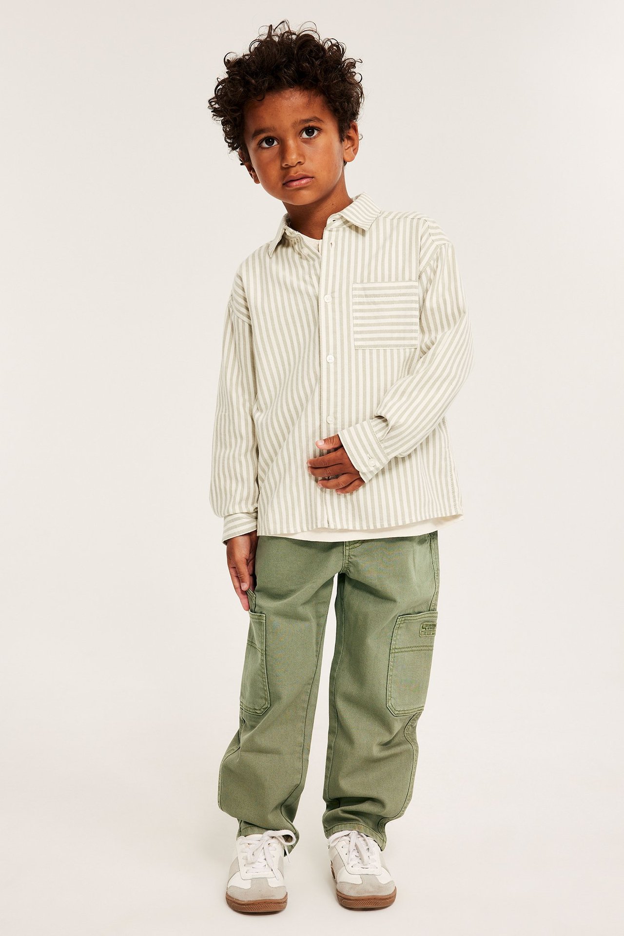 Widok z przodu dziecka w ciemnozielonych spodniach cargo, koszuli w paski i białych trampkach.