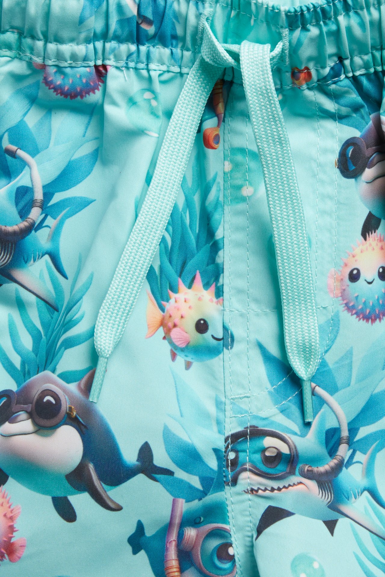 Nærbilde: Lyse turkise badeshorts for barn med haier og sjødyrmønster, samt snøring i midjen.