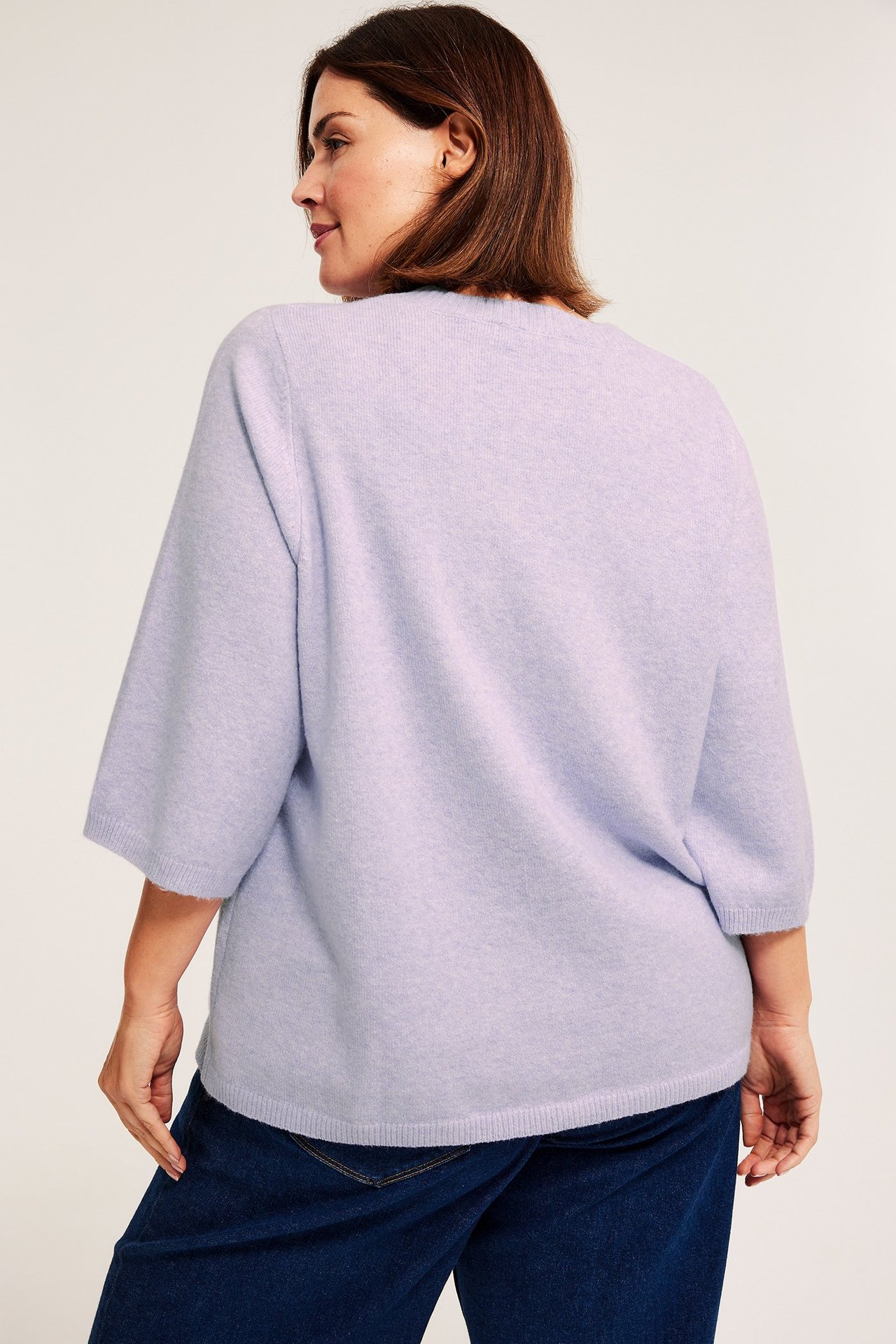Naisen takakuva vaaleanliilassa meleeratussa neulotussa t-paidassa 3/4-hihoilla ja tummansinisissä farkuissa.