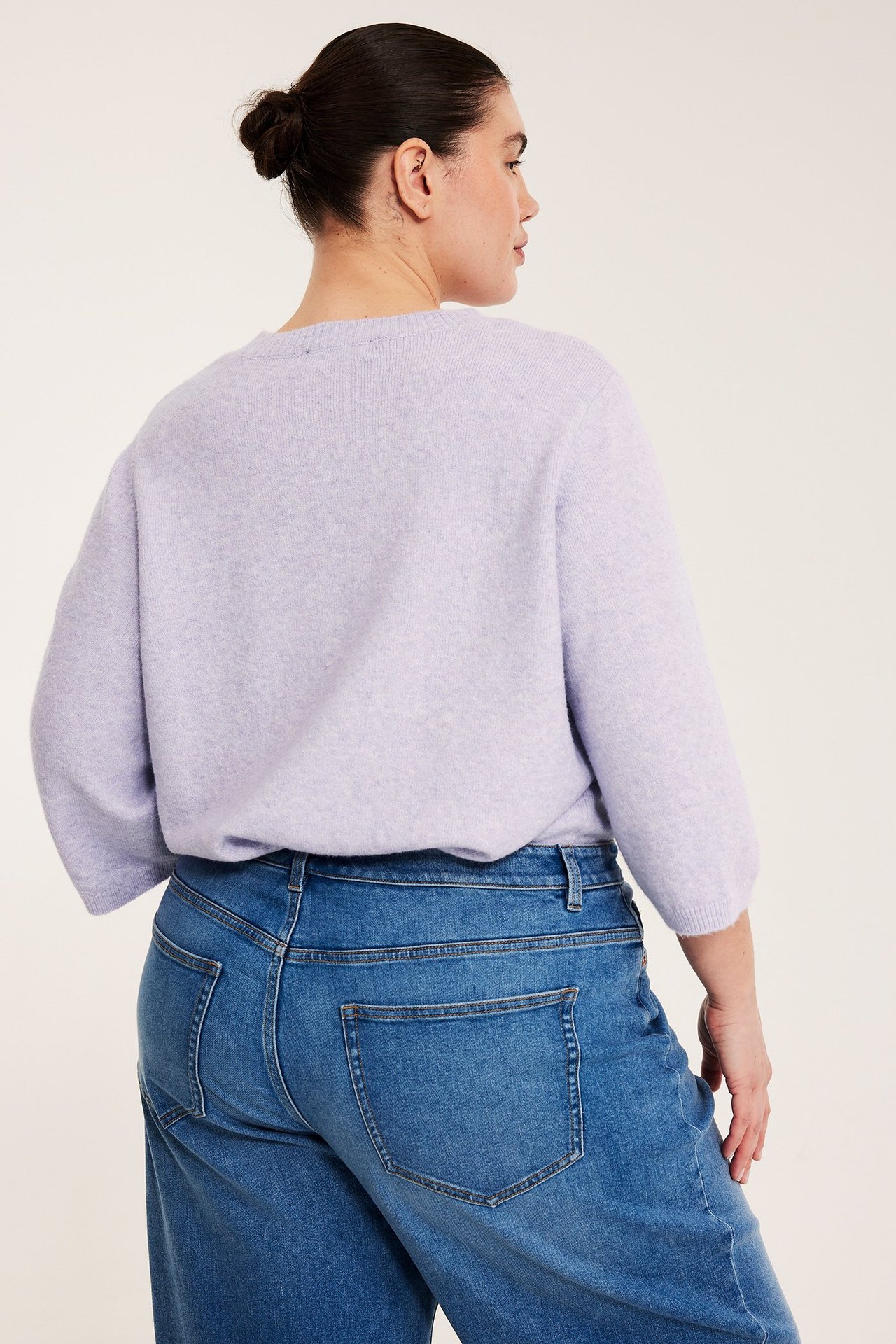 Tył kobiety w jasnofioletowej melanżowej dzianinowej koszulce z rękawem 3/4 i niebieskich jeansach.