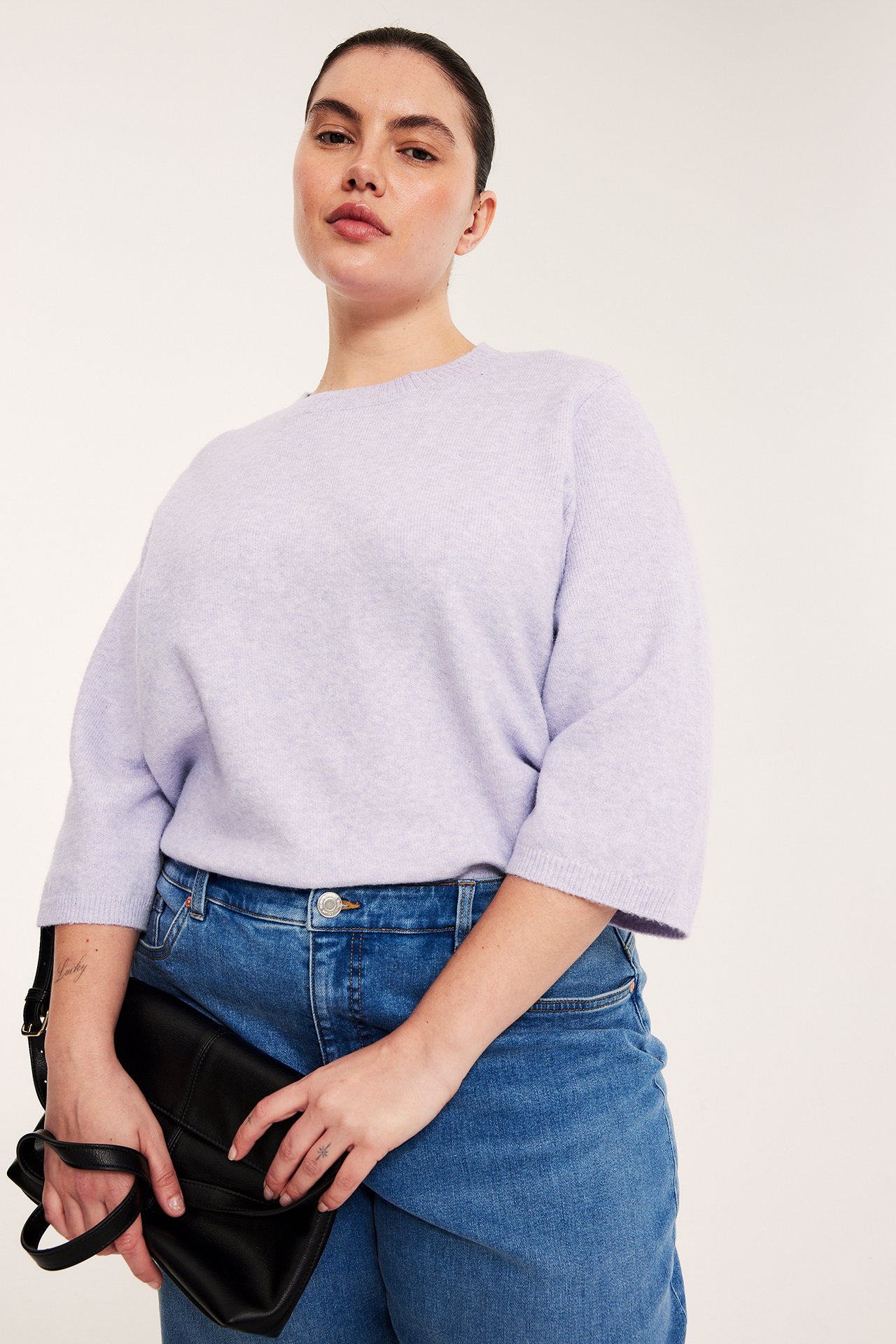 Nainen vaaleanliilassa meleeratussa neulotussa 3/4-hihaisessa t-paidassa, sinisissä farkuissa, pitäen mustaa käsilaukkua. Edestä.