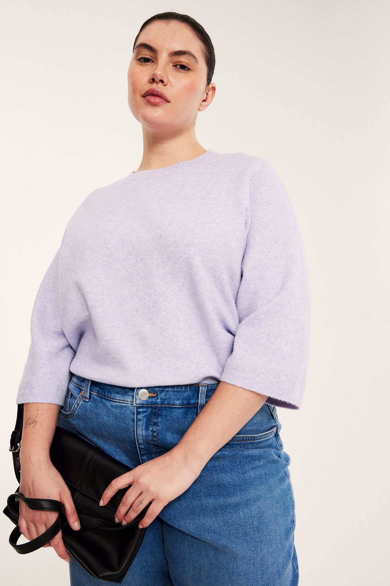 Kvinne i lys lilla melert strikket 3/4-ermet t-skjorte, blå jeans, holder svart håndveske. Frontvisning.