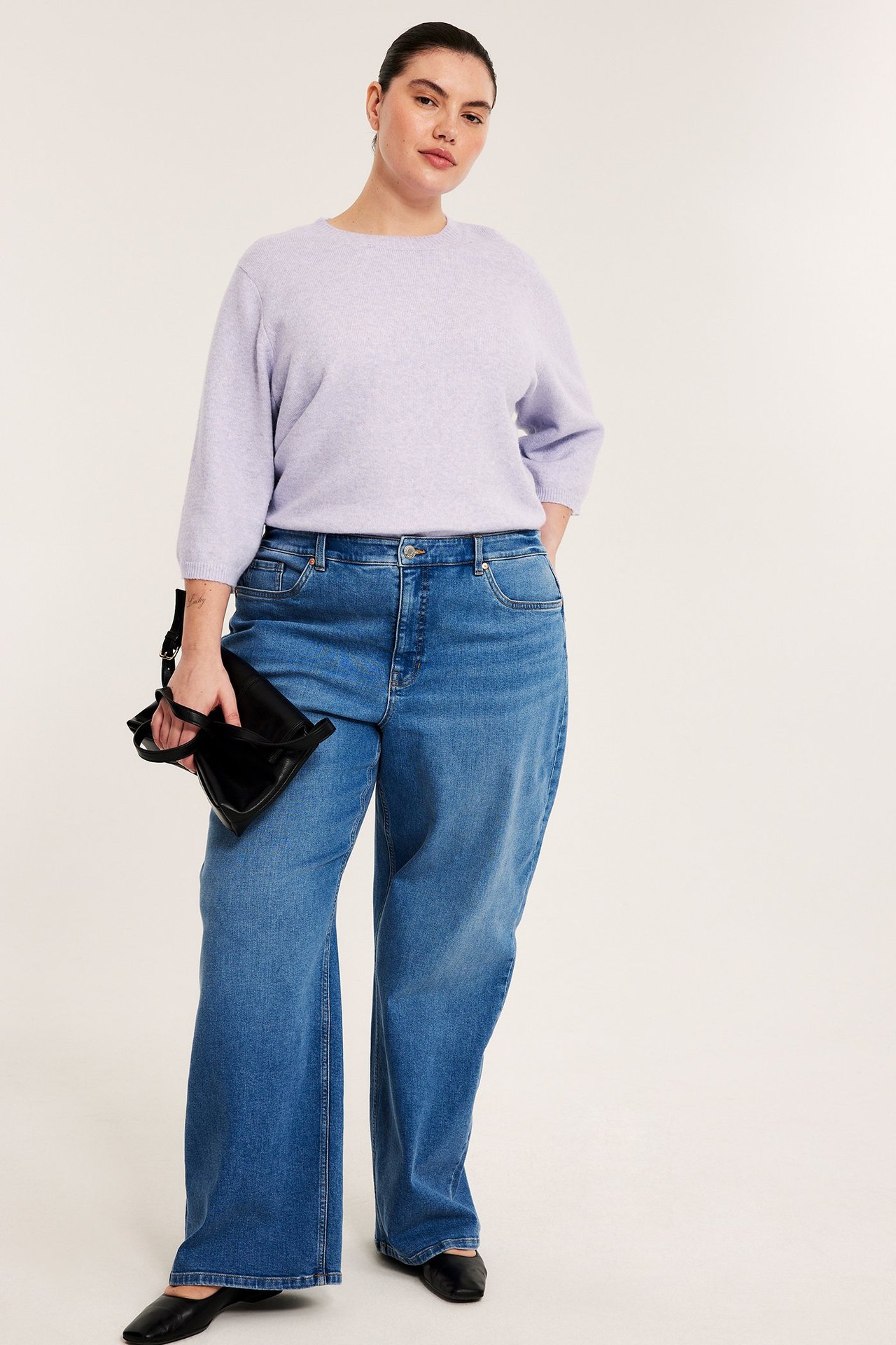 Forfra: Kvinne i lys lilla melert strikket 3/4-arm t-skjorte, blå vide jeans, holder en svart håndveske.