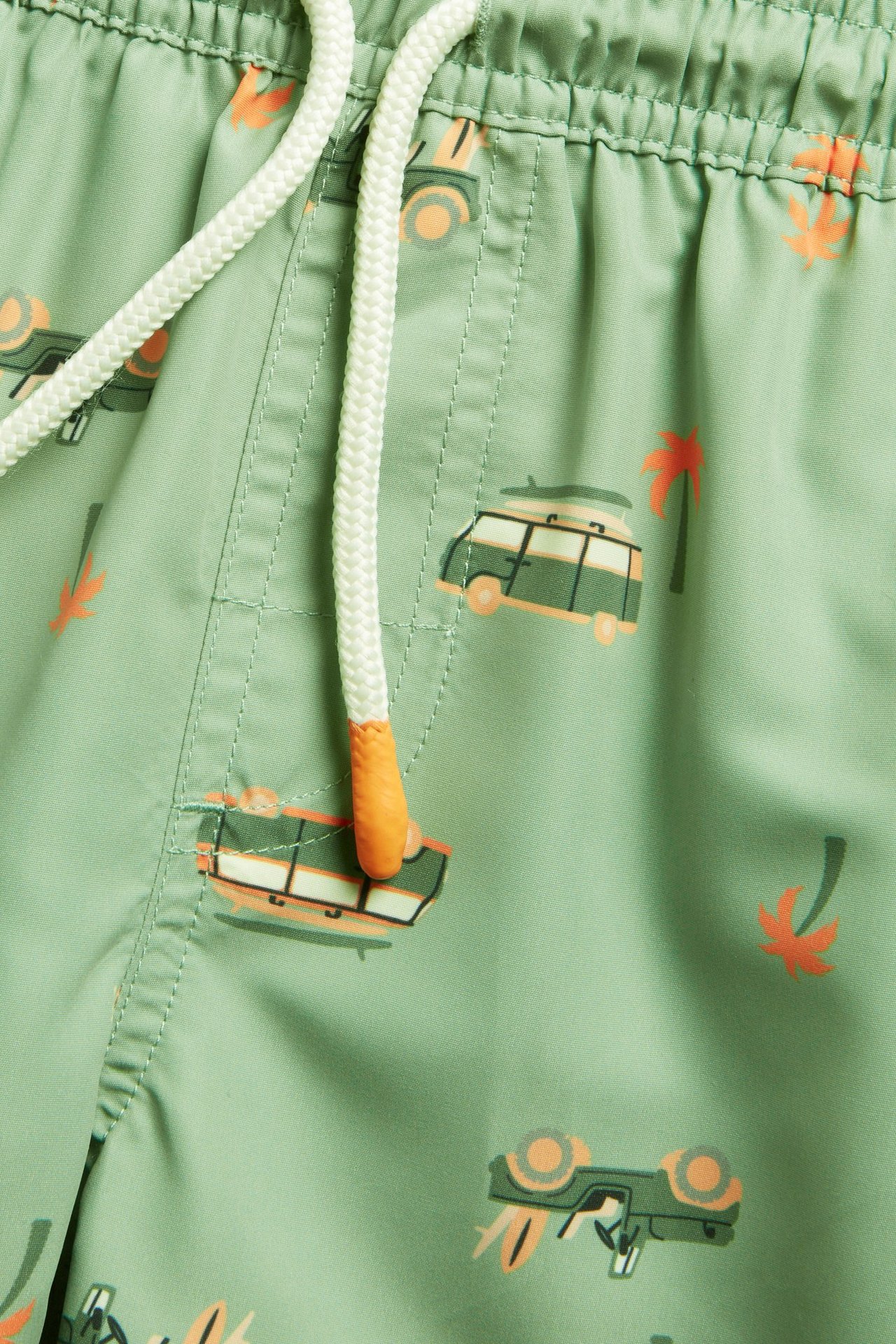 Lähikuva lasten vihreistä kuvioiduista uimahousuista, joissa pakettiauto- ja palmupuukuvio sekä valkoinen kiristysnyöri.