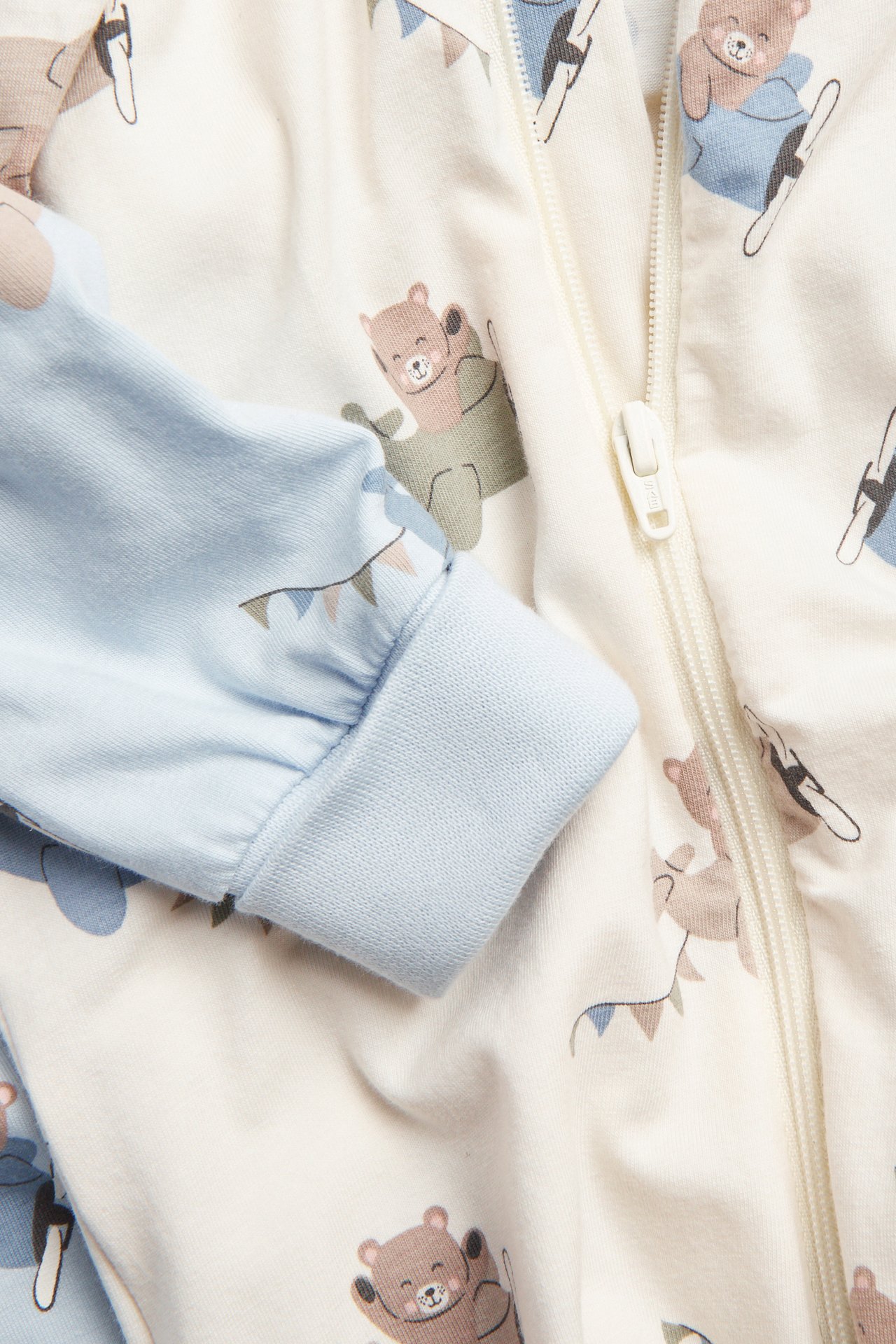 Lähikuva: luonnonvalkoinen vauvanhaalari karhu- ja lentokonekuviolla, valkoinen kaksisuuntainen vetoketju ja vaaleansiniset pidennetyt hihansuut.