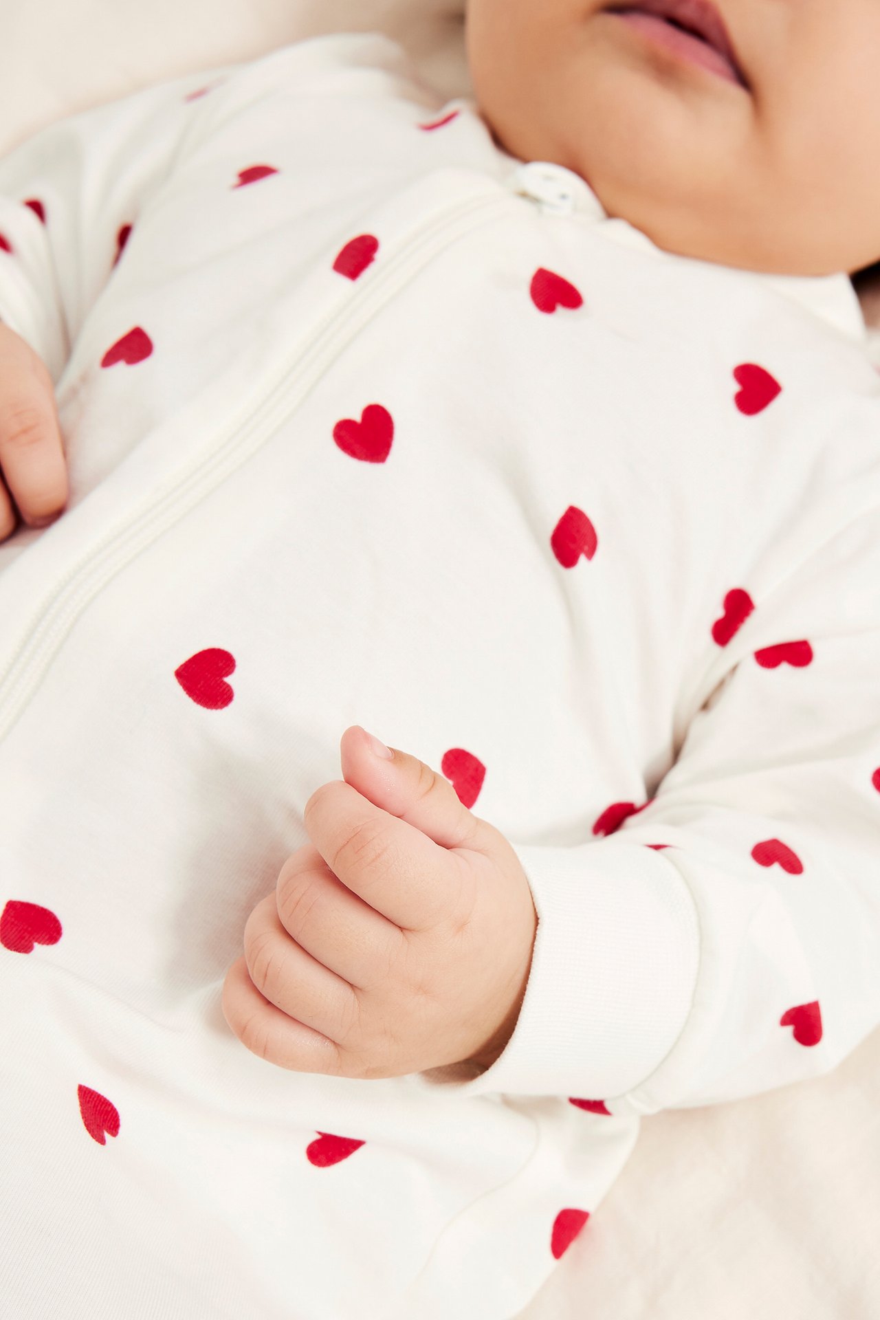 Närbild, framifrån, av en baby i offwhite pyjamas med rött hjärtmönster och dragkedja.
