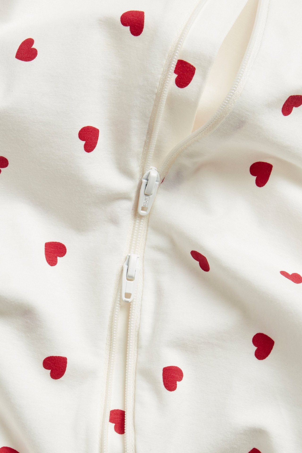 Lähikuva vauvan luonnonvalkoisista puuvillapyjamista, joissa punainen sydänkuvio ja valkoinen vetoketju.