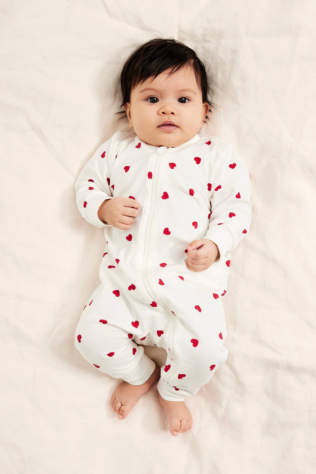Edestä, vauva yllään luonnonvalkoinen puuvillapyjama punaisella sydänkuviolla ja vetoketjulla edessä.