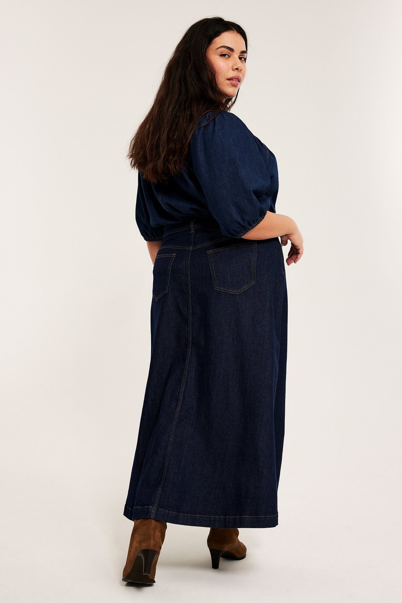 Rygg-sidovy av kvinna i mörkblå A-linje lång jeanskjol, matchande topp och bruna klackstövlar.
