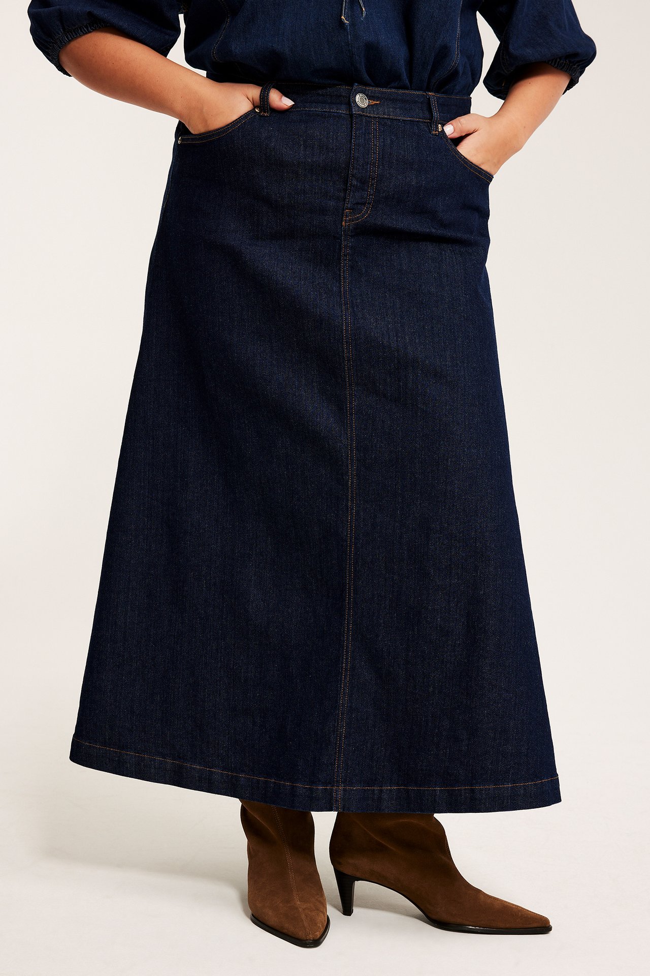 Frontvy av en kvinna i en mörkblå A-linje jeanskjol, en mörk topp och bruna mockakängor.